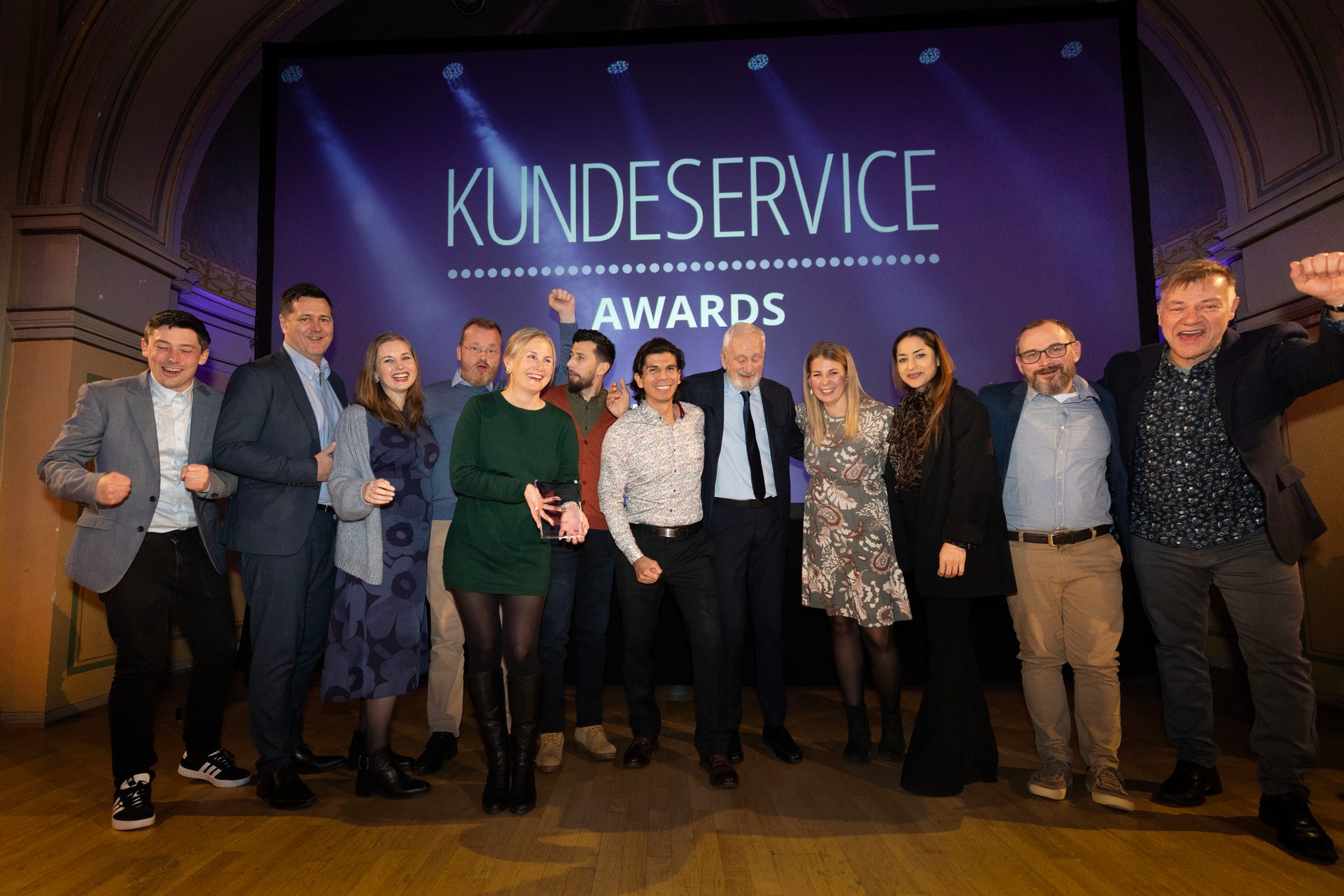Group of people celebrating on a stage in front of a screen displaying "KUNDESERVICE AWARDS".