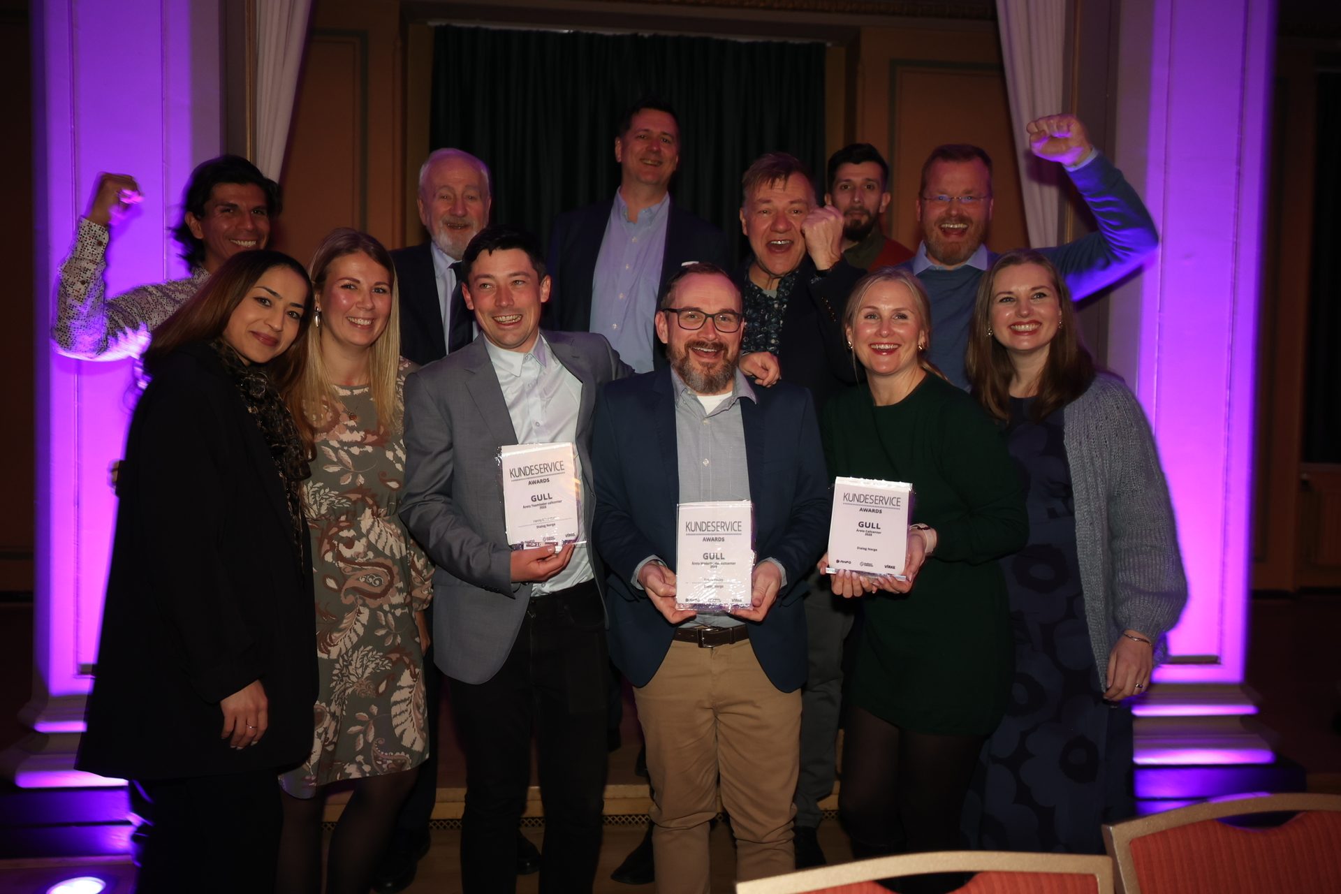 A group of smiling colleagues celebrating and holding 'KUNDESERVICE AWARDS GULL' trophies.