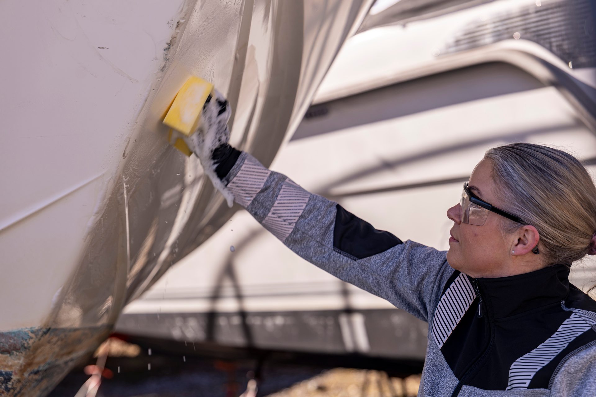 Woman wearing safety glasses and grey jacket scrubbing a white boat hull with a yellow sponge.