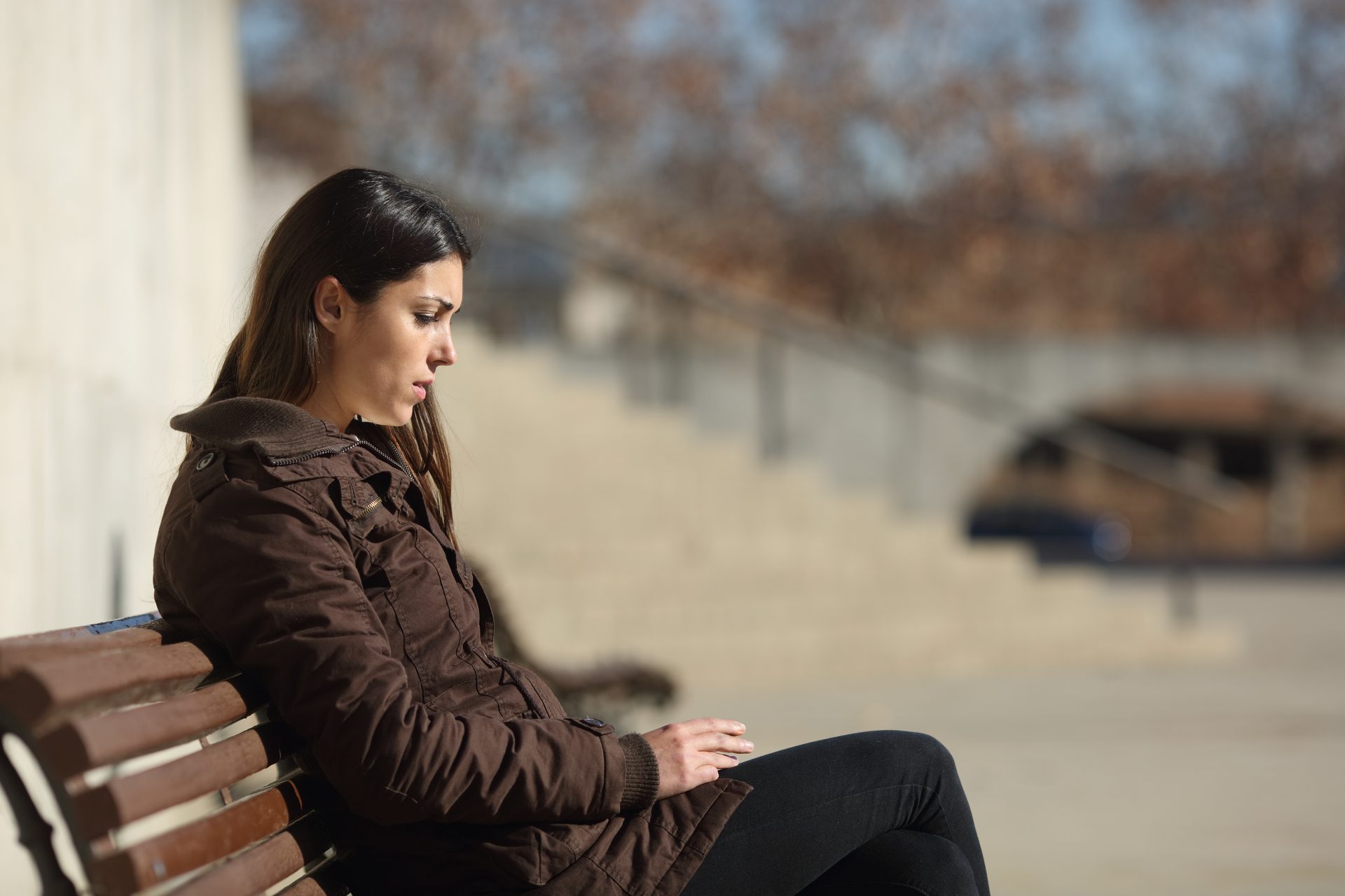 A young woman sits alone on a park bench, looking down with a sad or thoughtful expression.