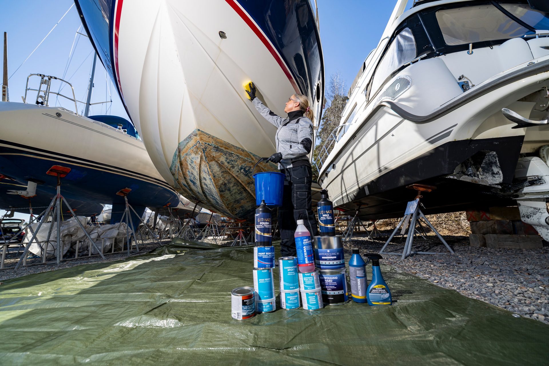 A woman cleans a boat's hull with a sponge, surrounded by various boat maintenance products on a green tarp.
