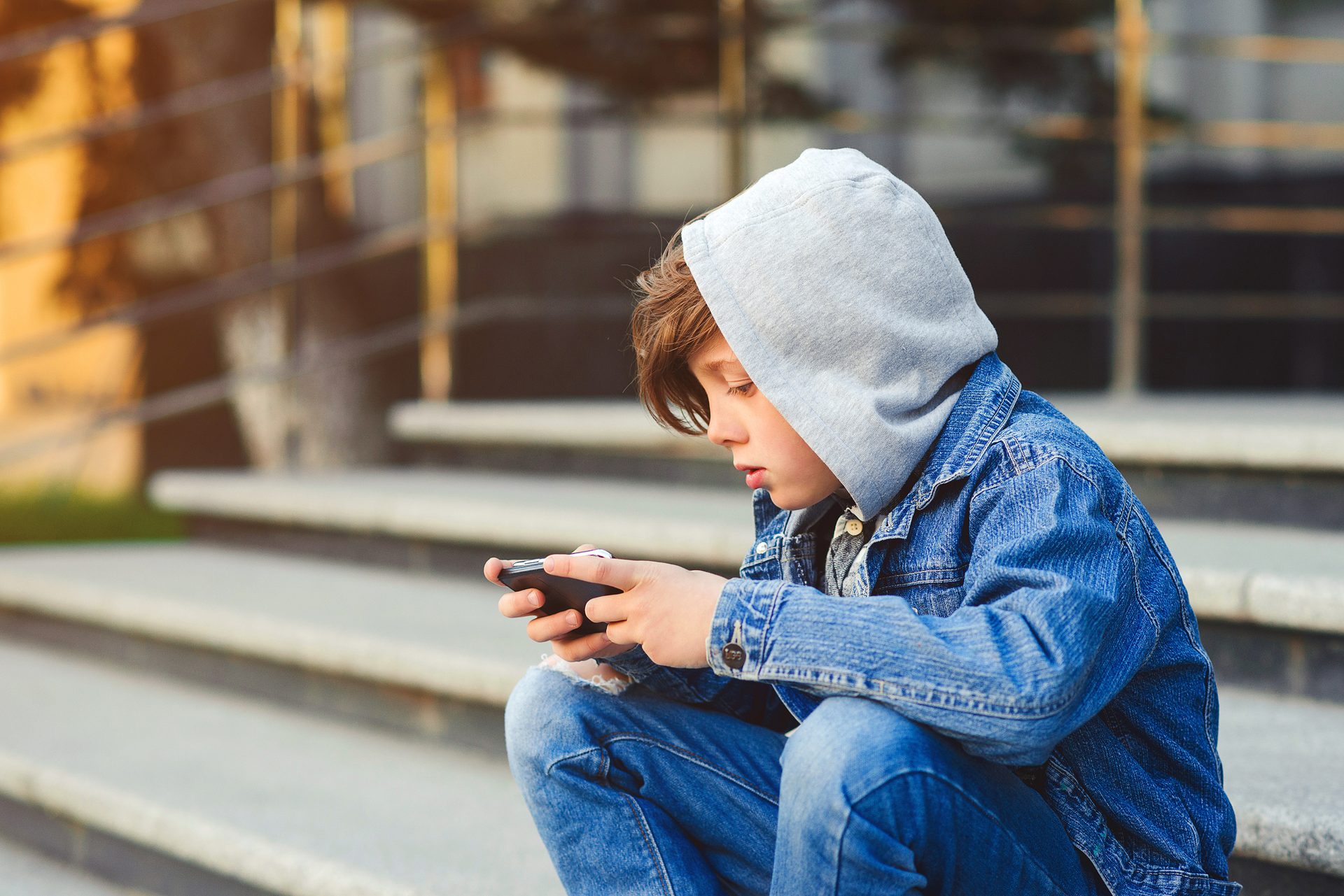 A boy in a gray hoodie and blue denim jacket sits on steps, absorbed in his smartphone.