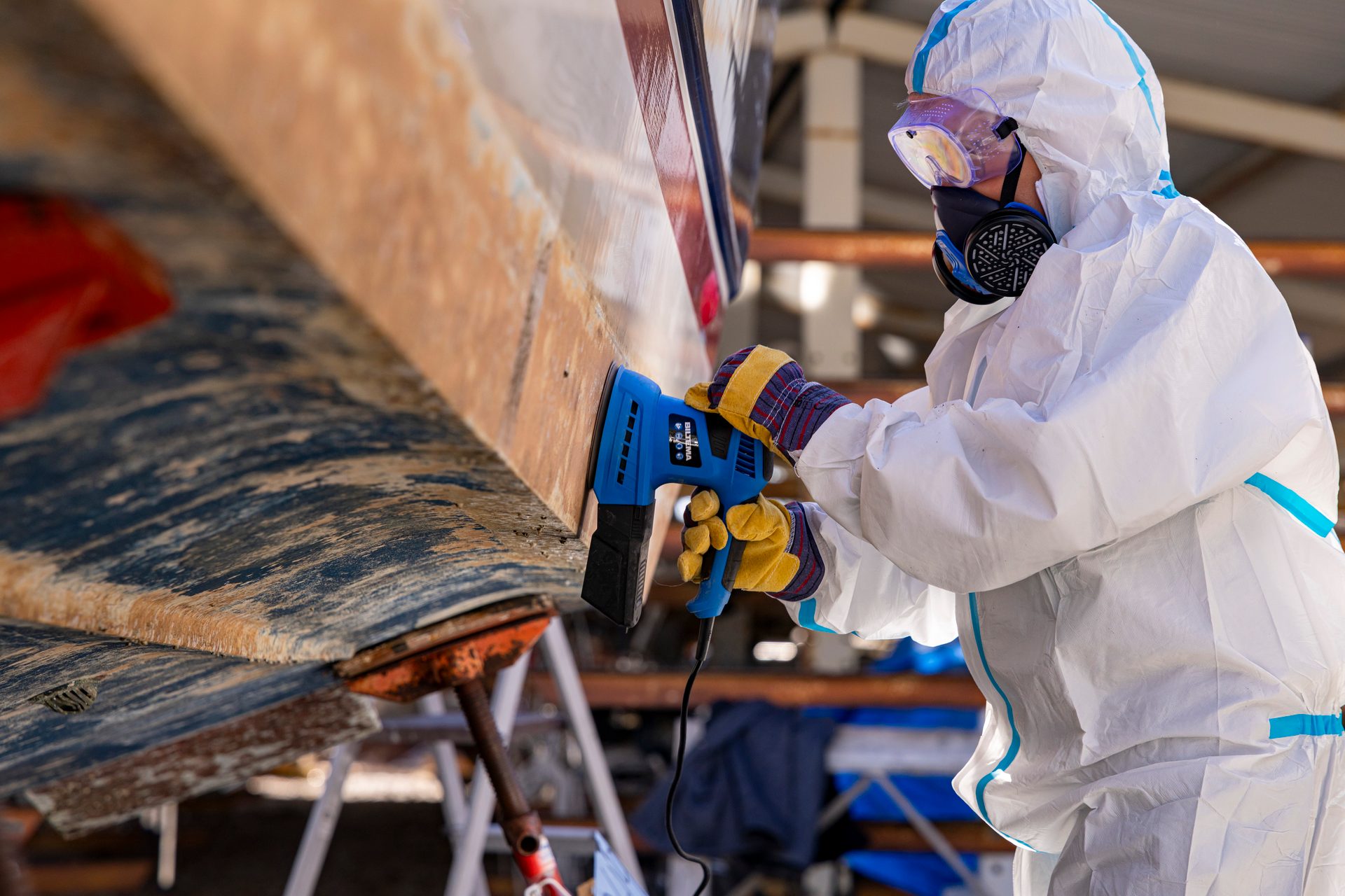 Person in protective suit sanding a boat hull with an electric sander, preparing for maintenance.