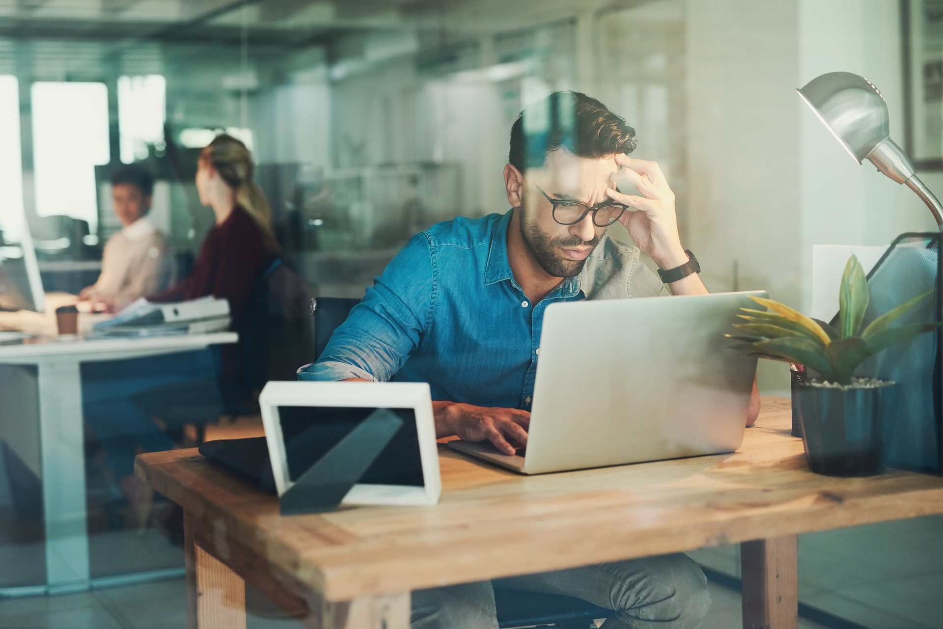 A man in glasses looks stressed while working intensely on a laptop at a desk in a modern office.