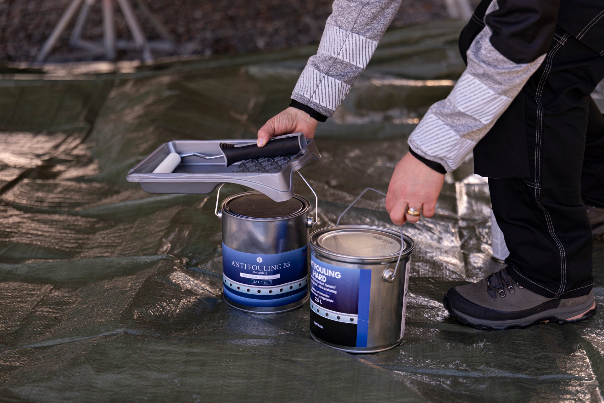 A person's hands holding a paint roller tray over two cans of anti-fouling boat paint on a tarp.