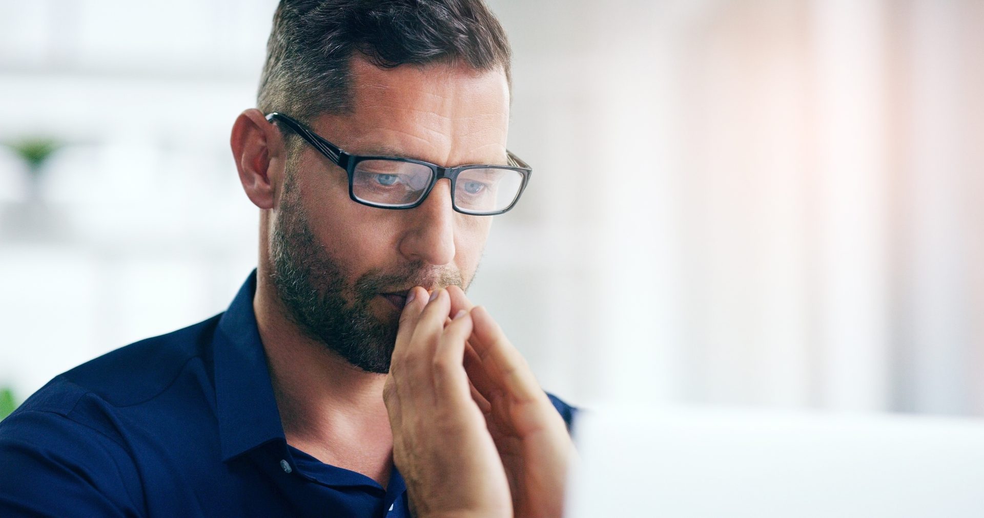 A man with glasses and a beard, wearing a blue shirt, looks down intently, hands clasped in thought.