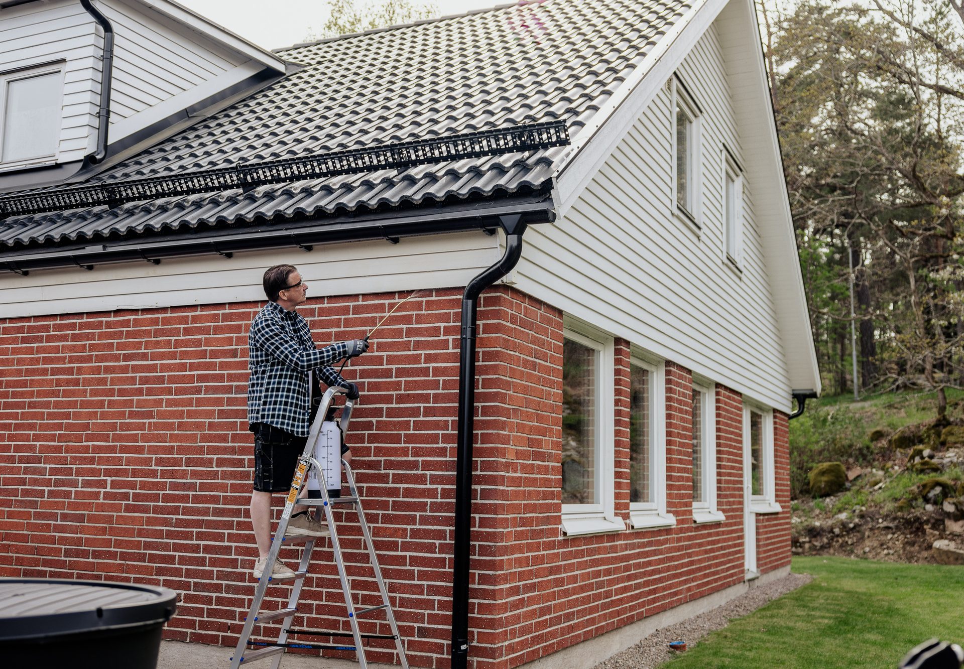 Man on a ladder spraying the gutter of a brick house with a white gabled roof.