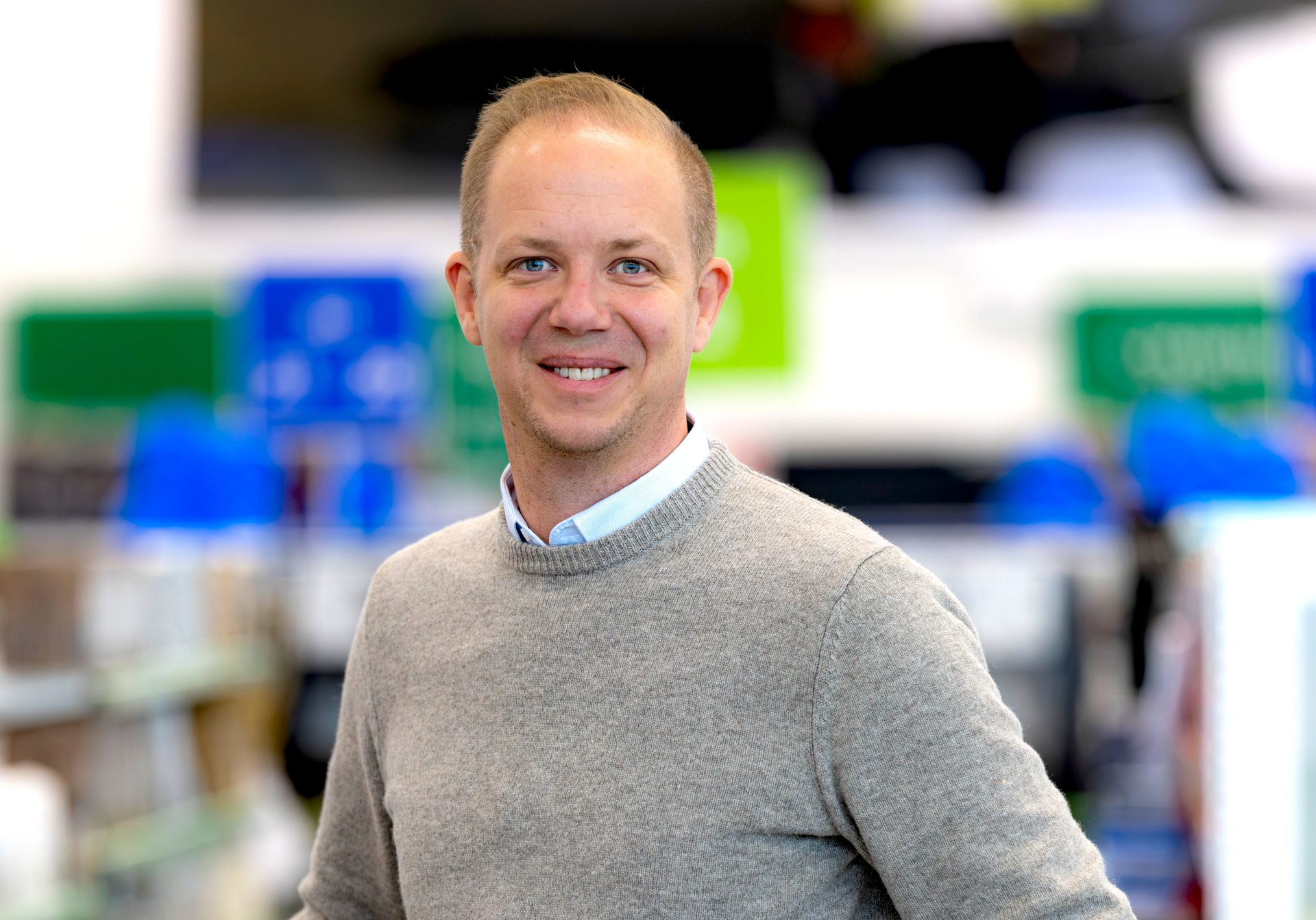 Smiling man in a gray sweater looks at the camera, against a blurred, colorful background.