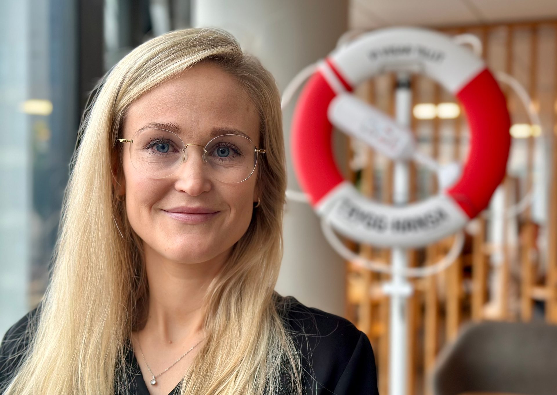 A blonde woman in glasses smiles at the camera, a red and white lifebuoy is blurred in the background.