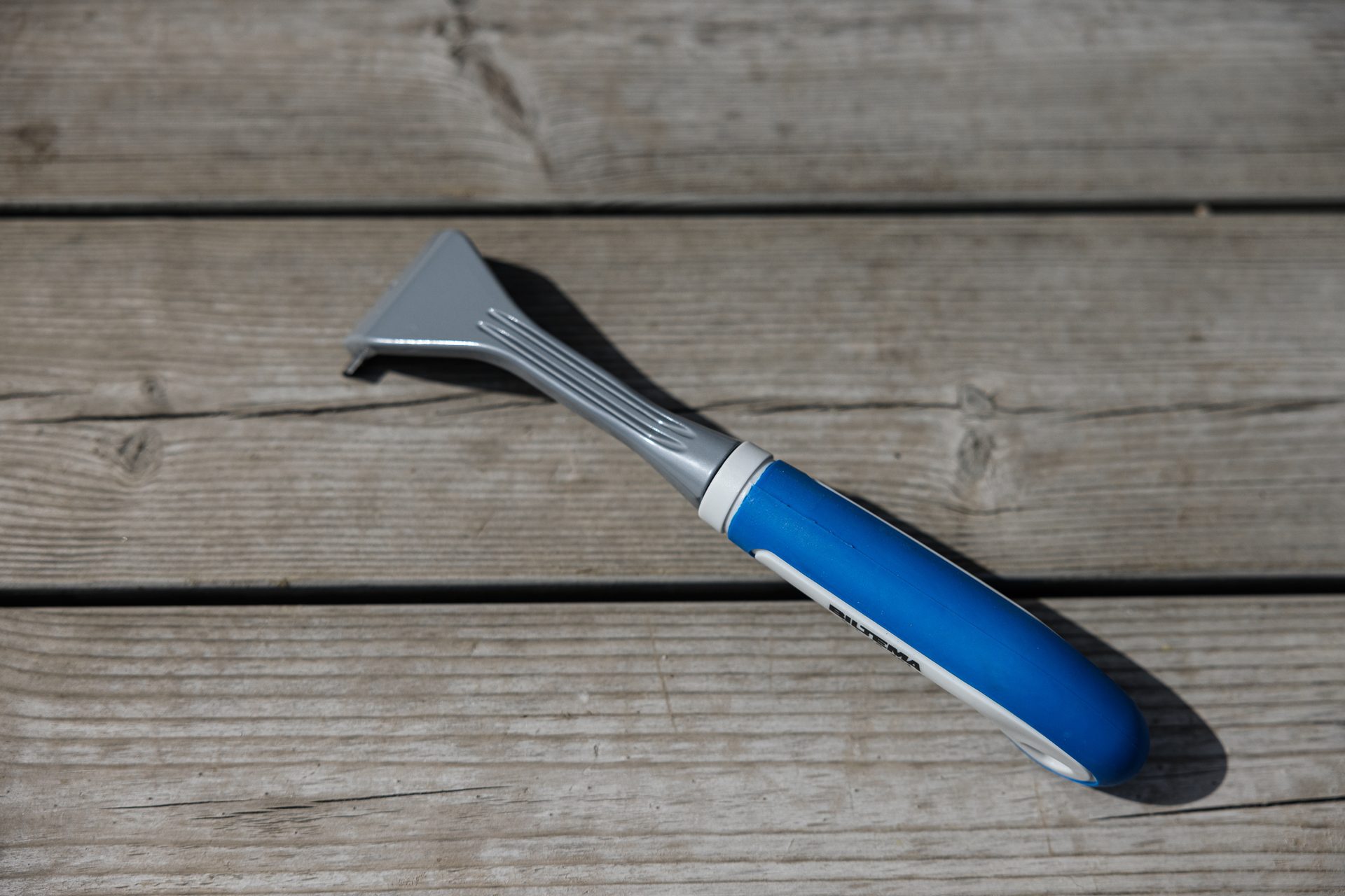 A blue and gray ice scraper rests diagonally on a weathered wooden deck surface.