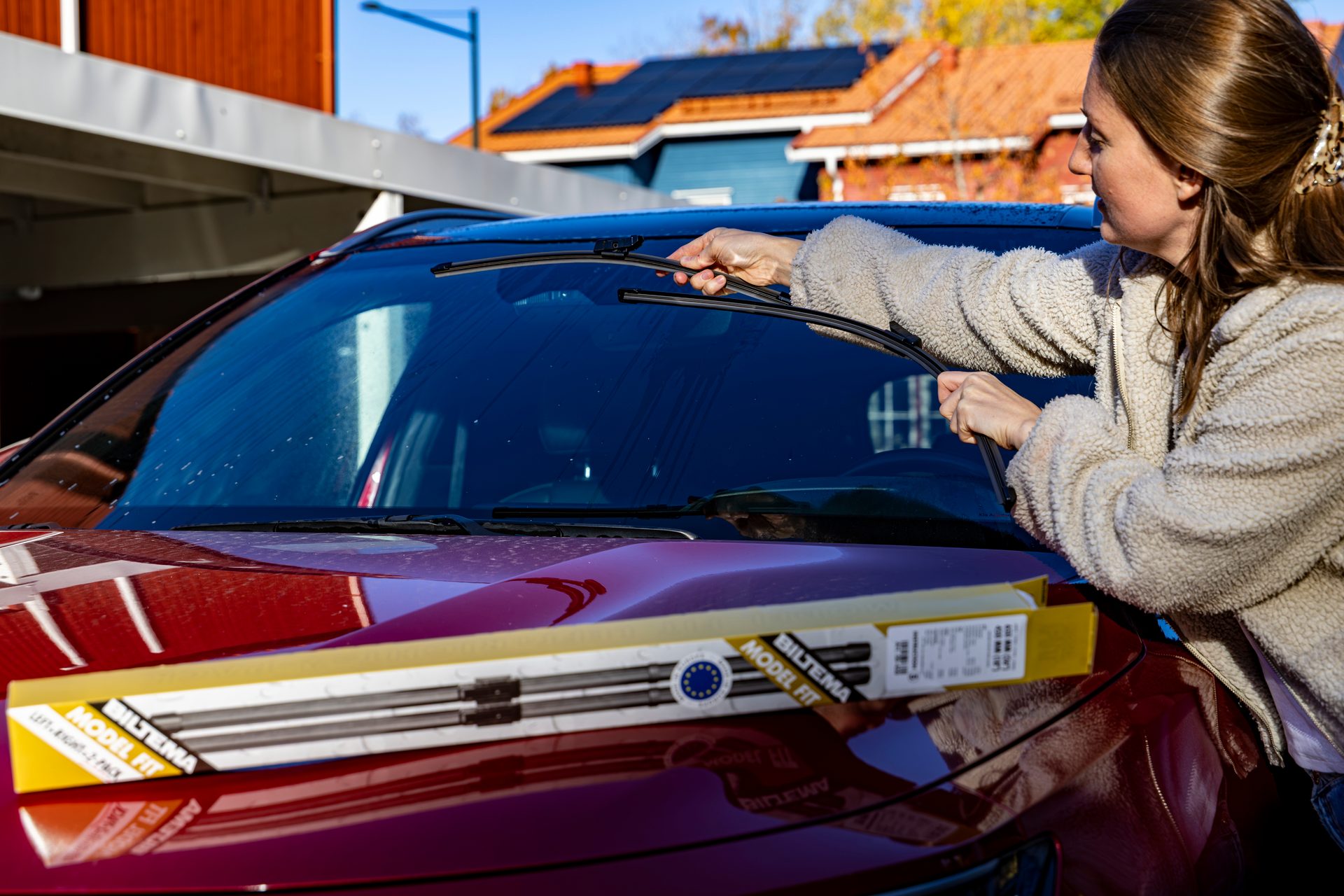 Woman changing windshield wipers on a red car, with a new wiper box on the hood.