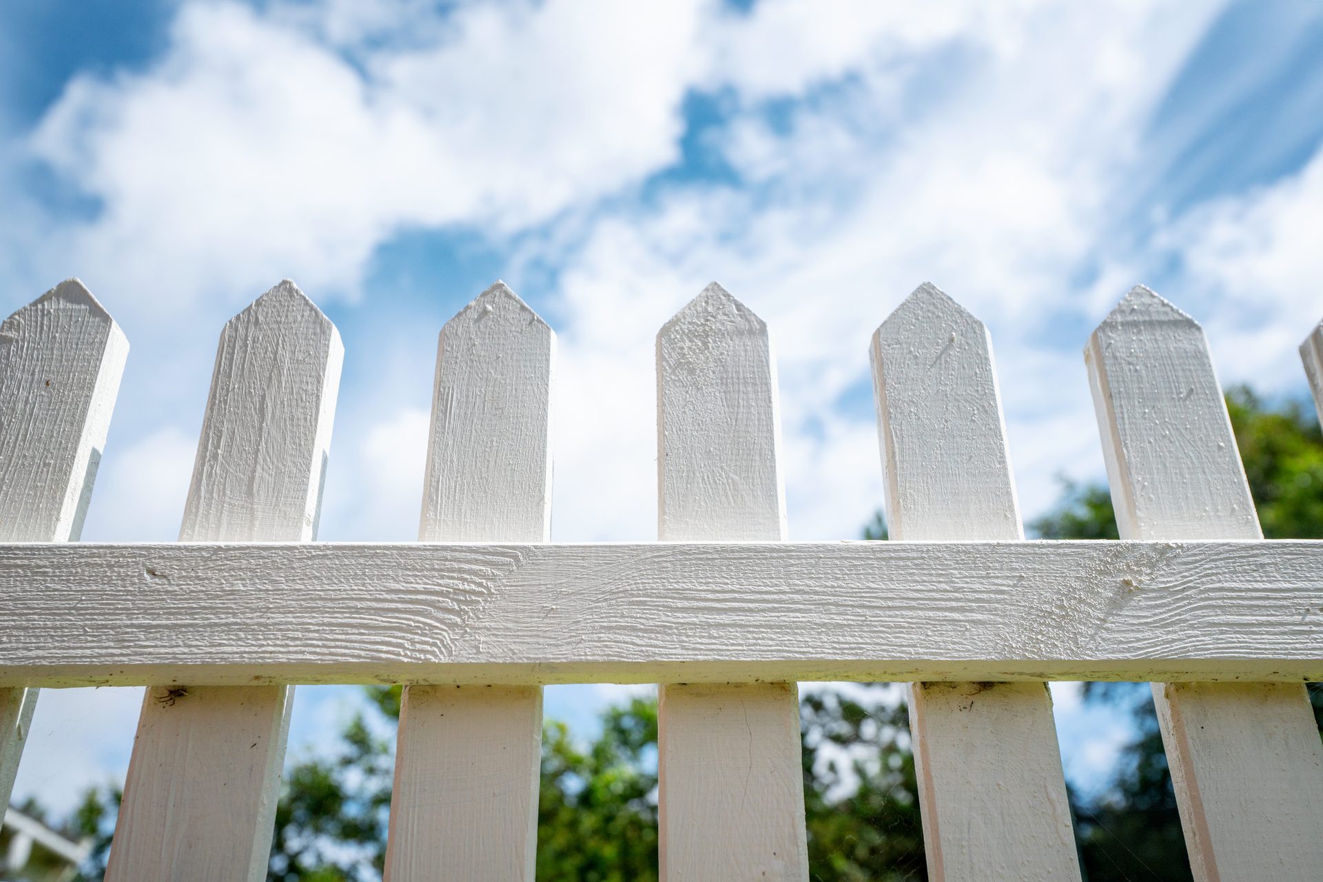 A low-angle view of a white picket fence against a bright blue sky with white clouds.
