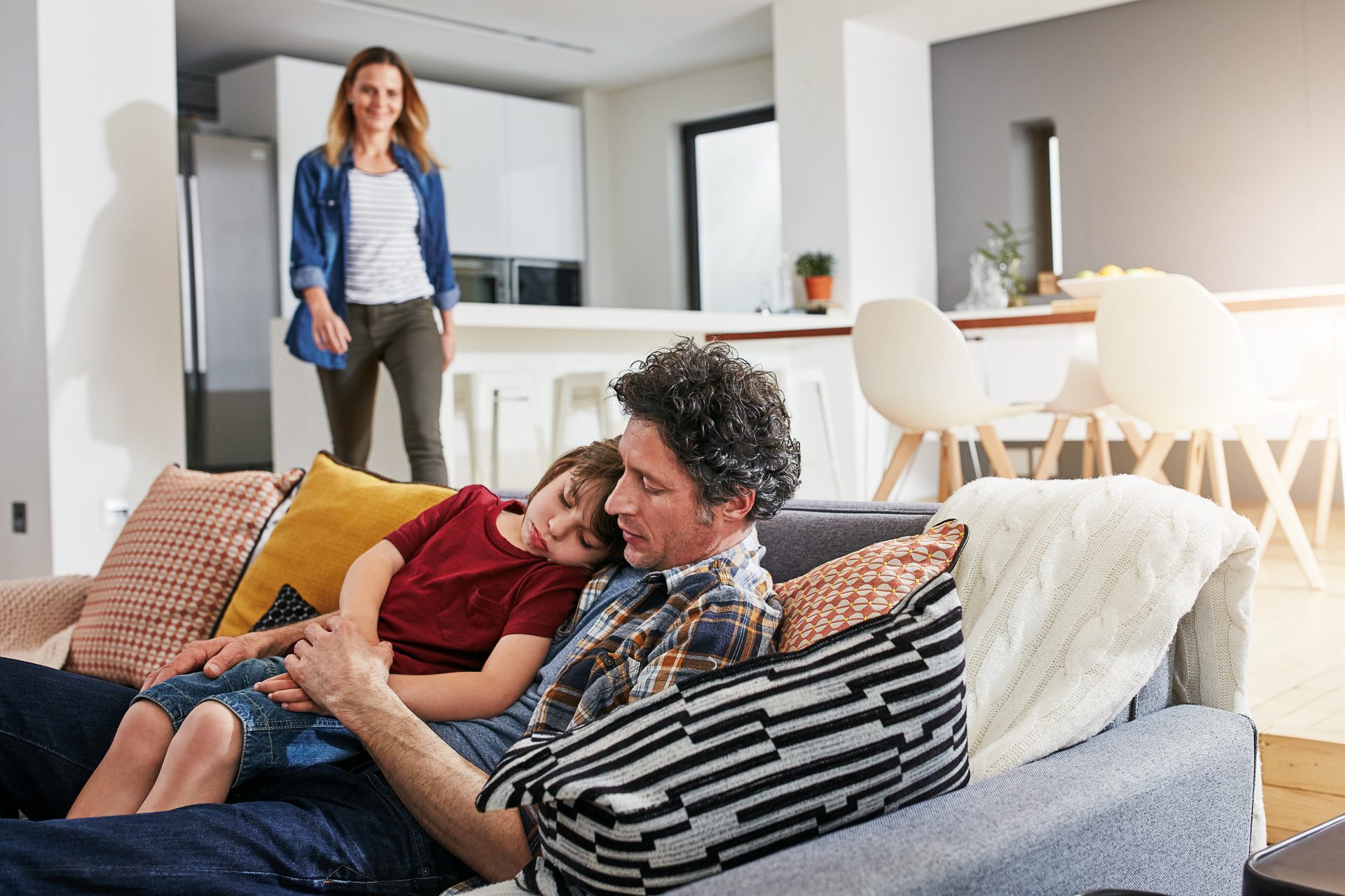 A family scene with a father and sleeping child on a couch, and a mother standing in the background.