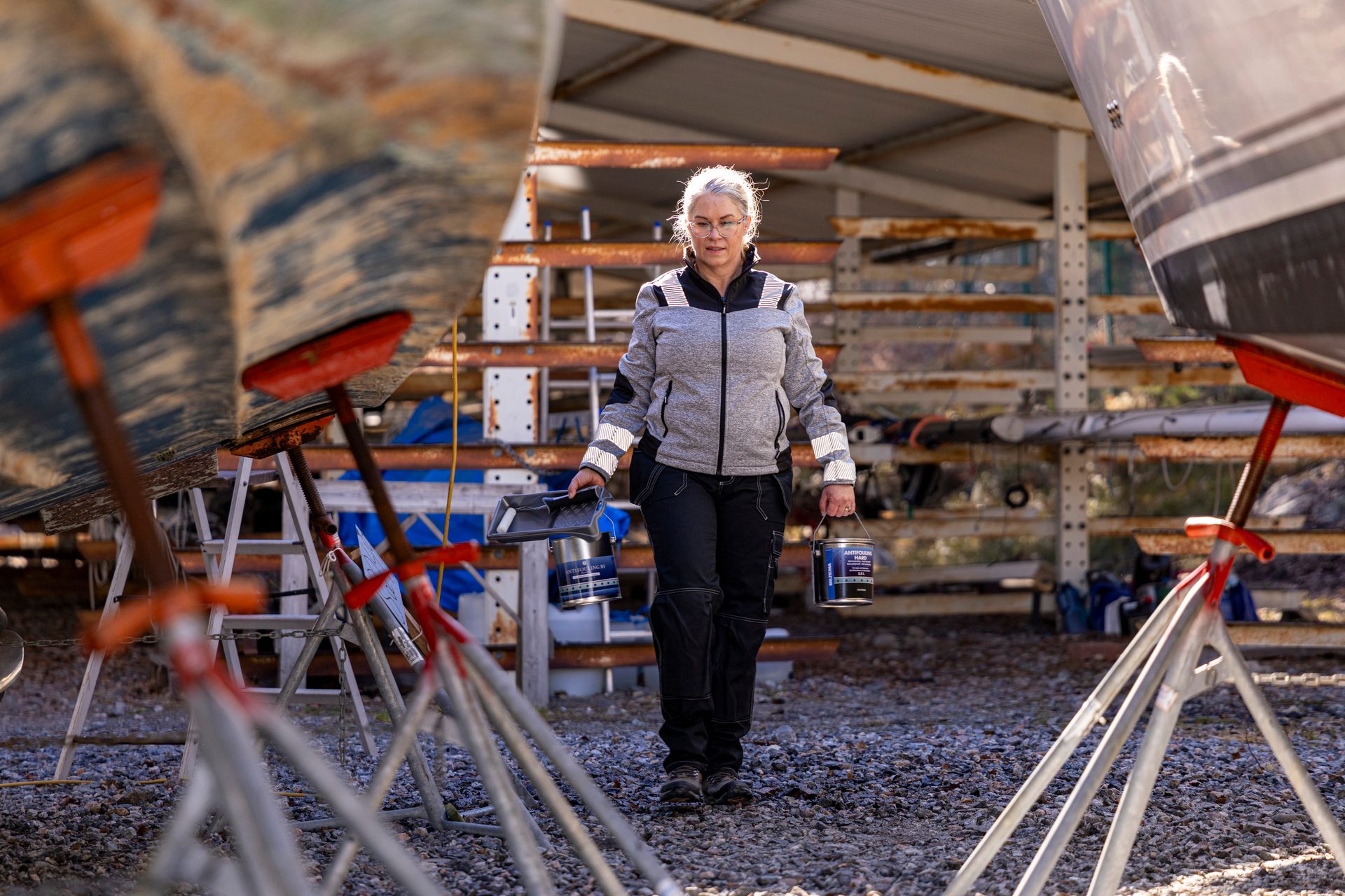 Woman carries paint cans and a tray, walking between boats in a shipyard, ready to work.