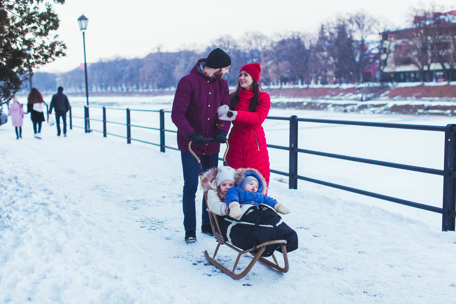 Parents pull two children in a sled on a snowy path by a frozen river, smiling.