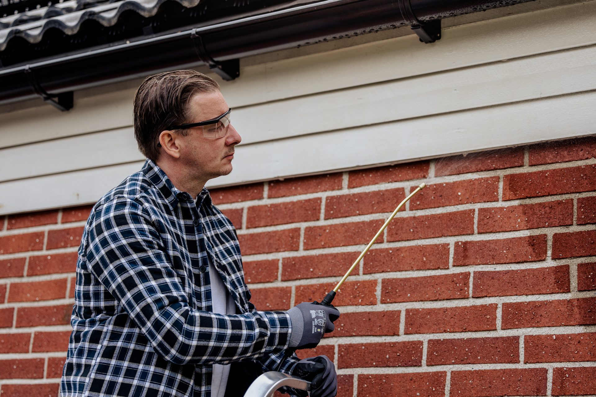 Man in safety glasses and plaid shirt sprays liquid onto a brick wall, likely for pest control or cleaning.