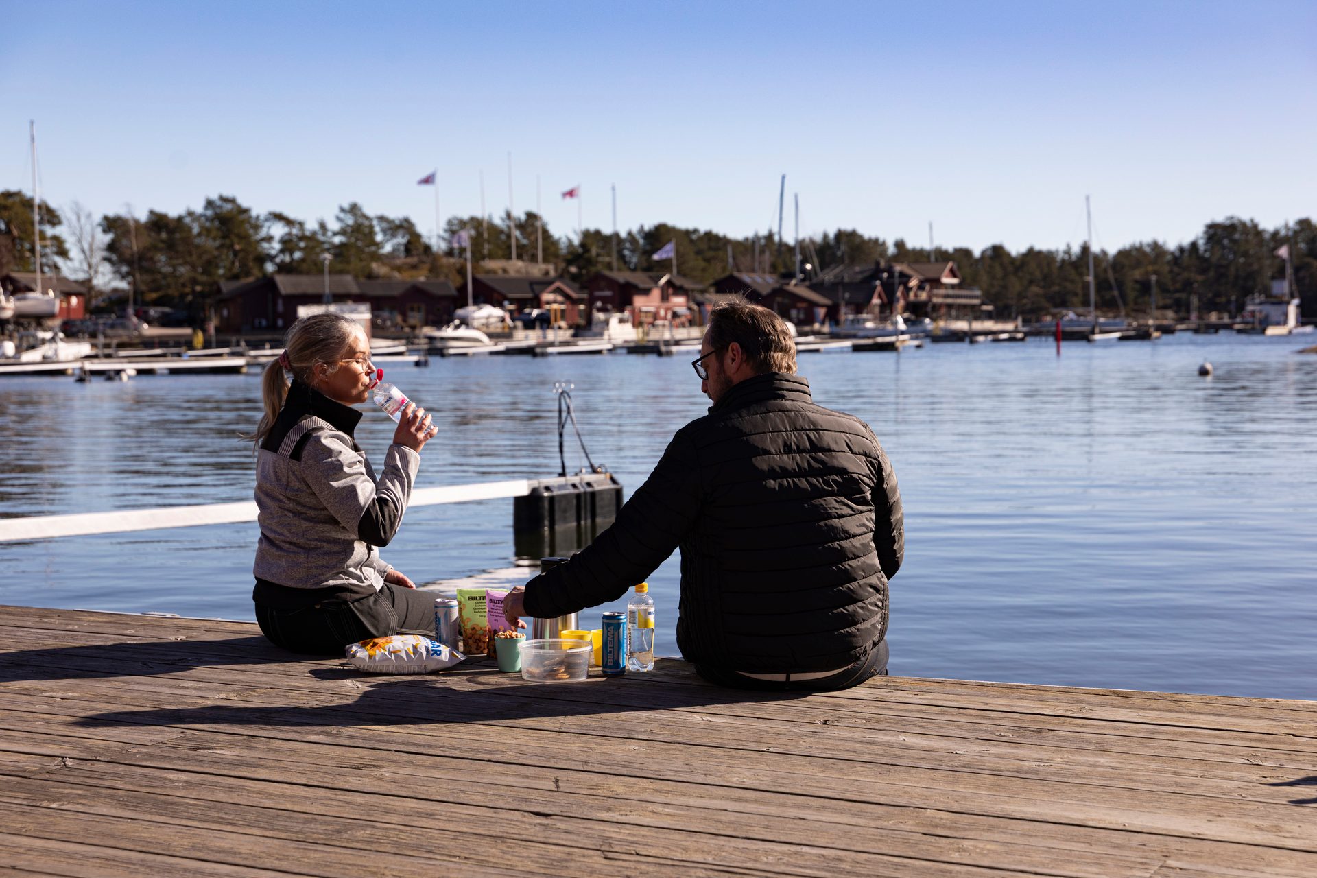 A couple sits on a wooden dock by a lake, enjoying a picnic with snacks and drinks on a sunny day.