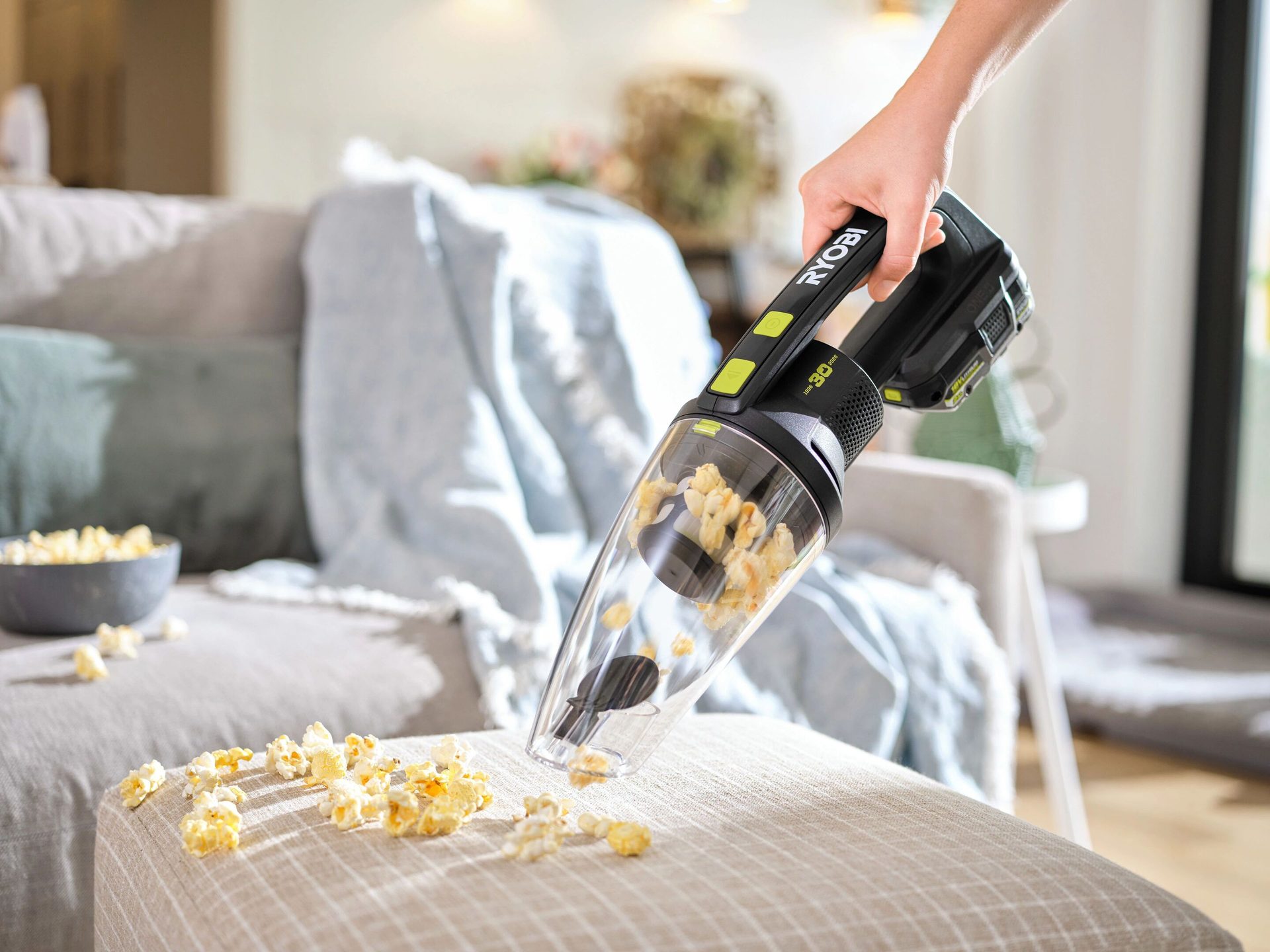 A hand holding a Ryobi handheld vacuum cleaning spilled popcorn from an ottoman in a living room.