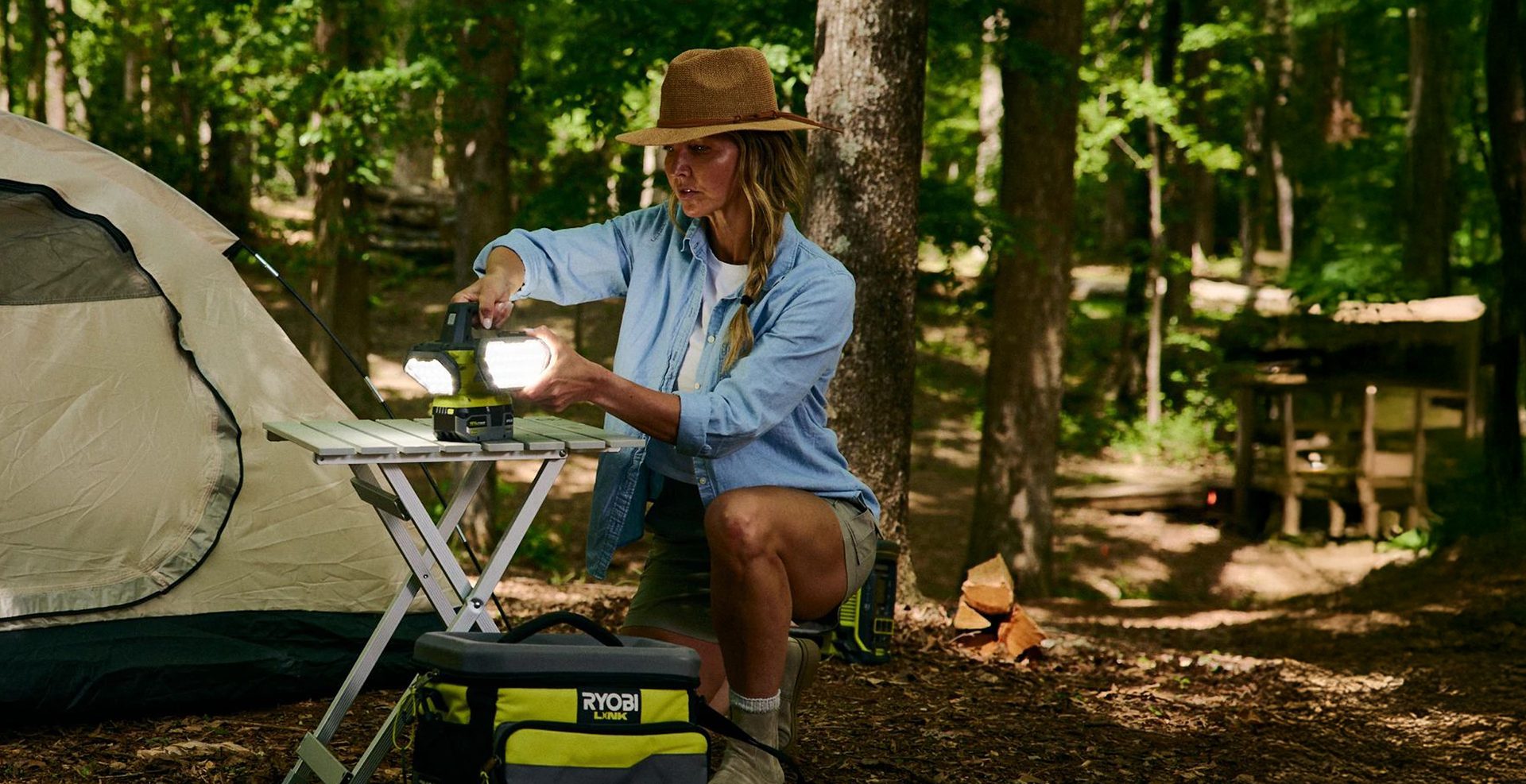 Woman adjusting a bright lamp on a table at a campsite with a tent and trees.