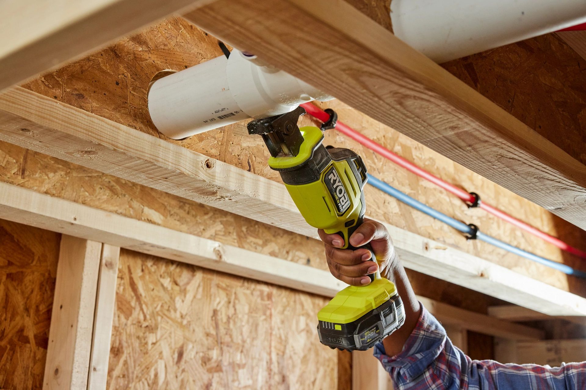 A person uses a Ryobi reciprocating saw to cut a white pipe near wooden beams and PEX plumbing.