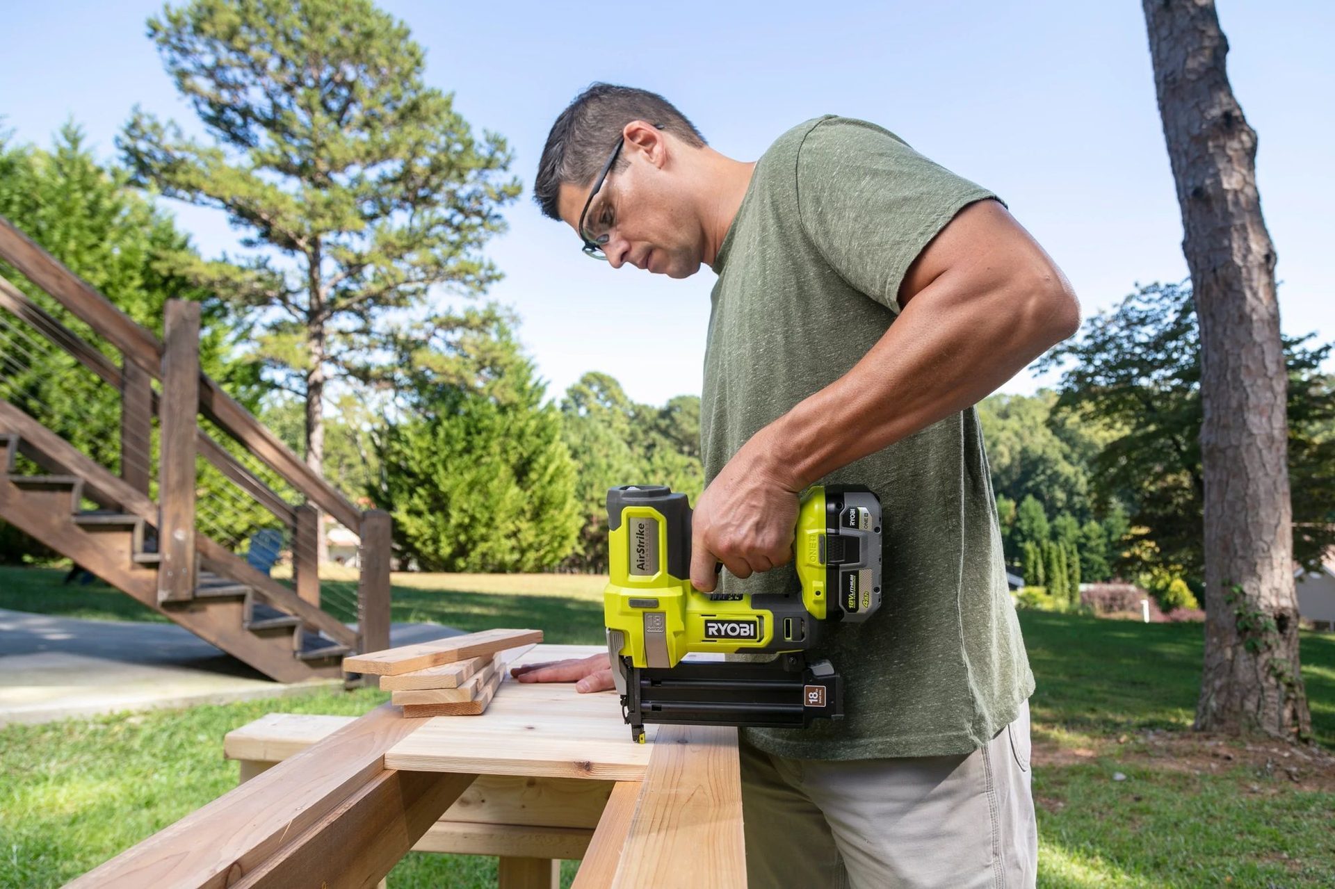 Man in safety glasses using a yellow Ryobi nail gun on wood planks outdoors.