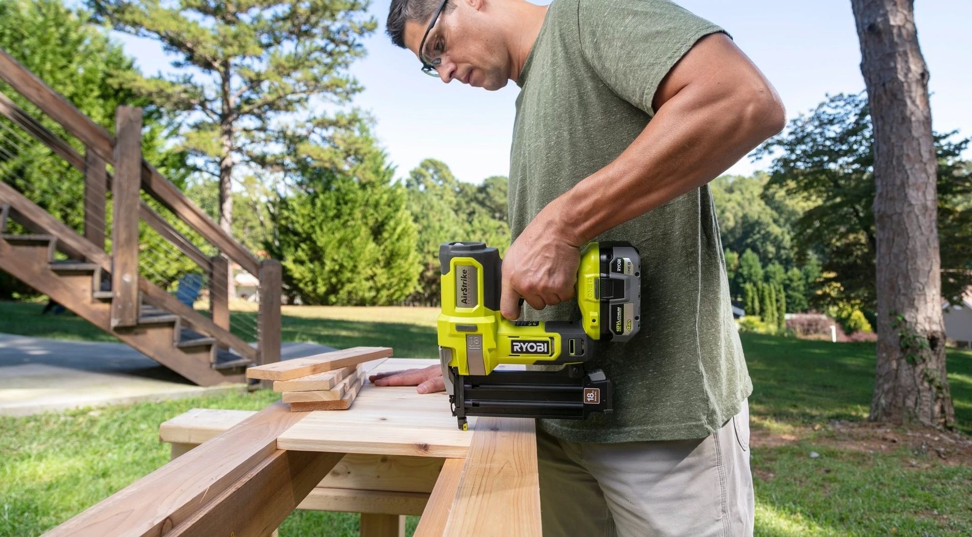 Man in safety glasses using a Ryobi Airstrike nail gun to secure wood outdoors.