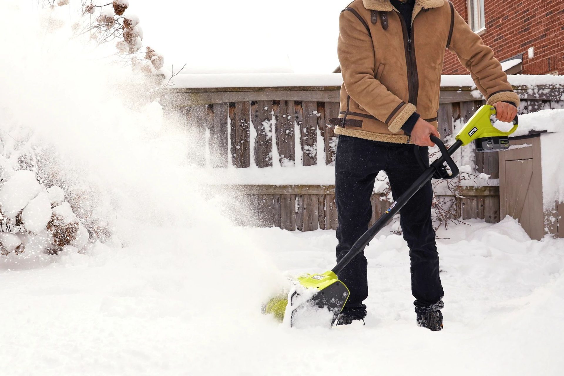 A person in a brown jacket uses an electric snow shovel to clear snow in a snowy backyard.