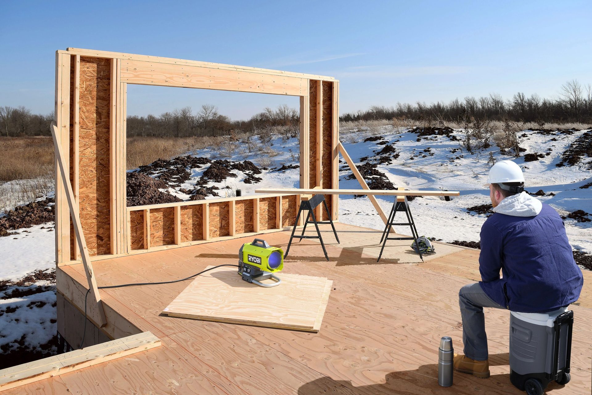 A worker sits on a cooler near a partially framed wall at a snowy construction site with a RYOBI fan.
