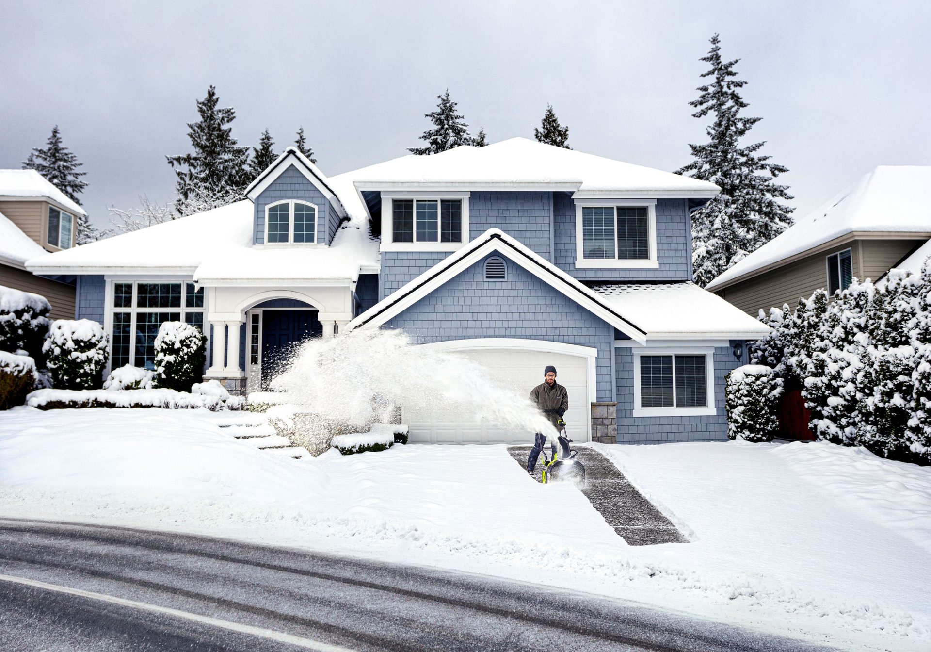 Man snow blowing a snowy driveway in front of a blue house during winter.