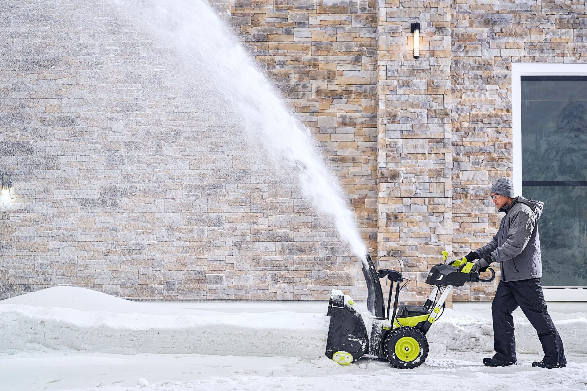 A man uses a snow blower, throwing snow, in front of a stone house.