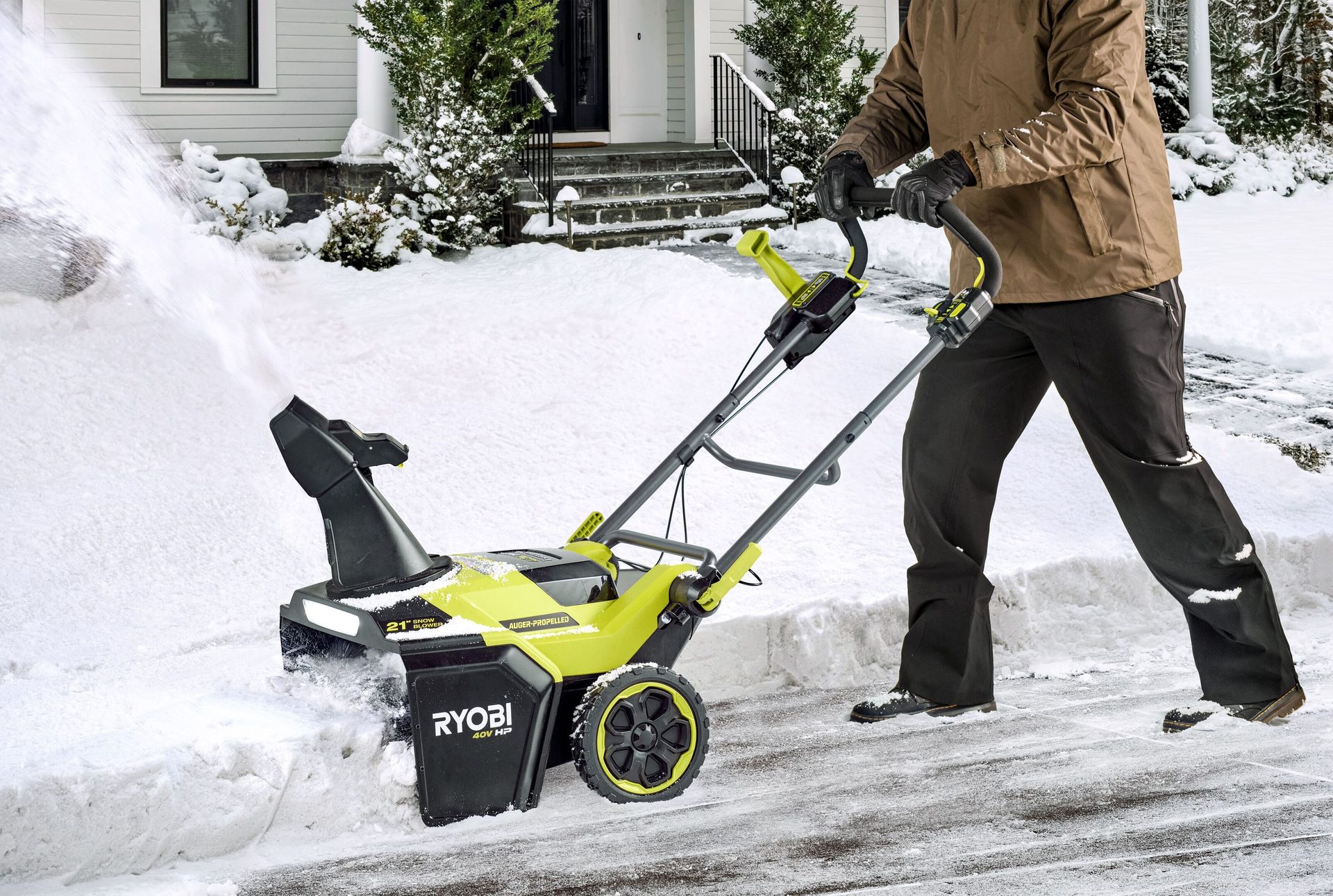 Man using a green Ryobi snow blower to clear snow from a path in front of a house.