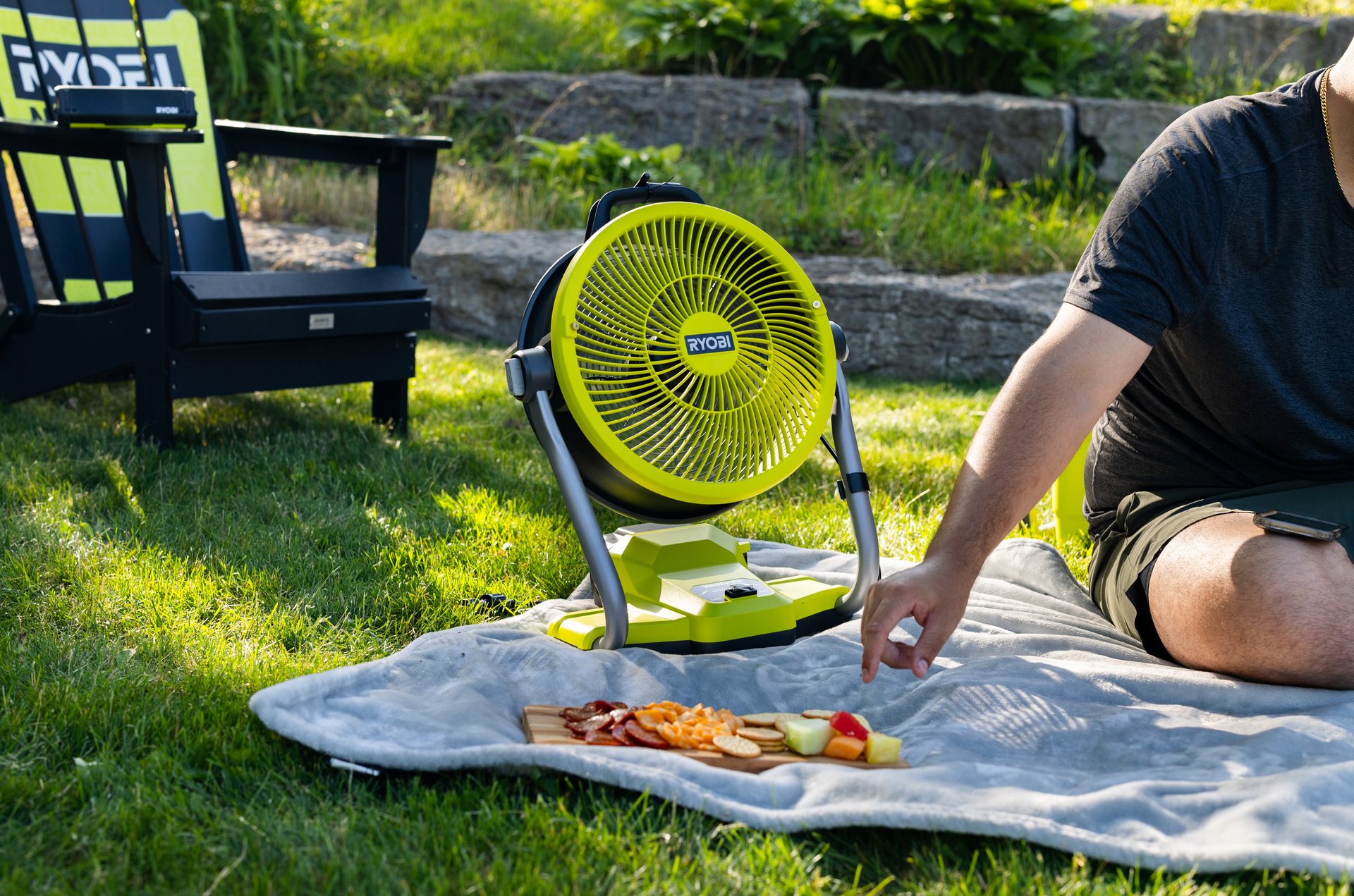 Person picnicking with food, a bright green Ryobi fan, and a branded chair on grass.