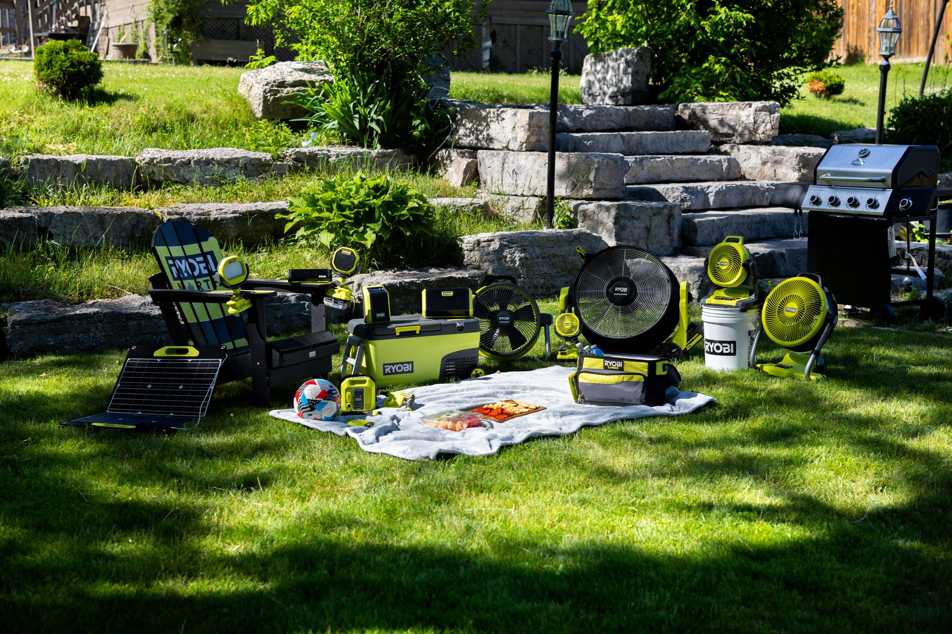 Ryobi fans, cooler, grill, chair, and picnic setup on a sunny lawn.