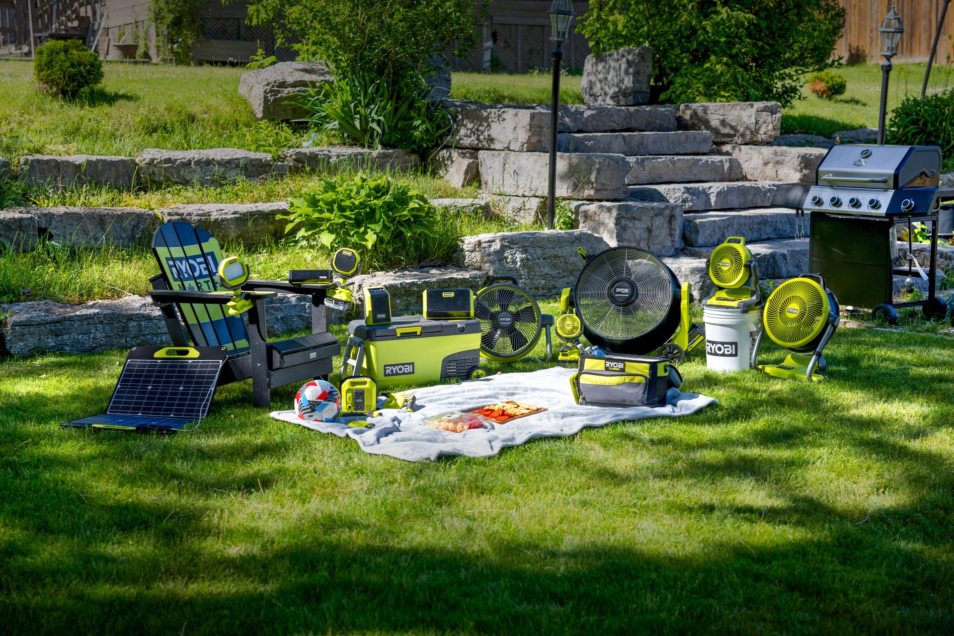 Ryobi outdoor living products: fans, cooler, solar panel, and grill on a sunny lawn.