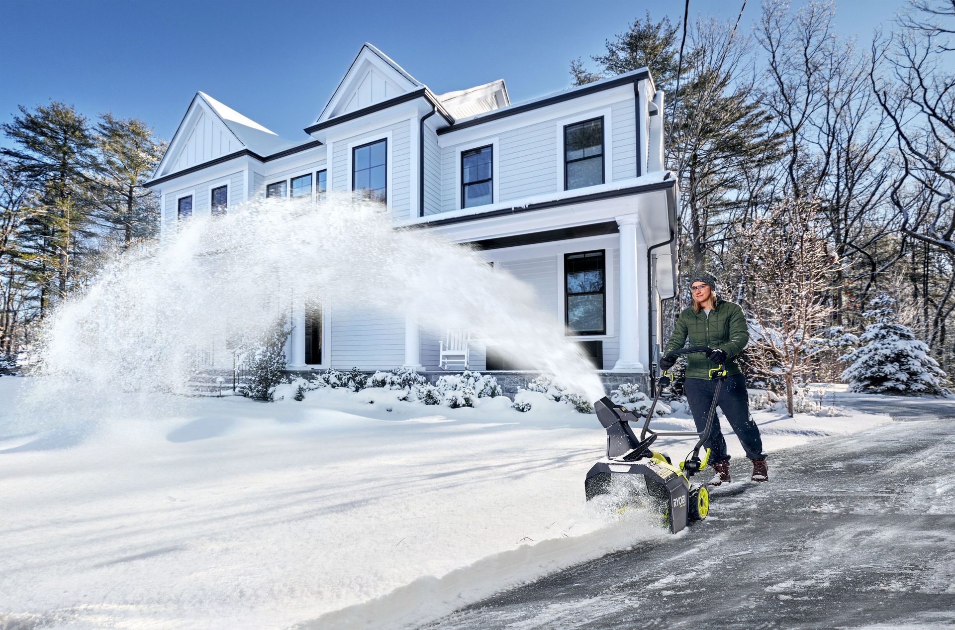 Man snow blowing a driveway with a snowblower in front of a large house.