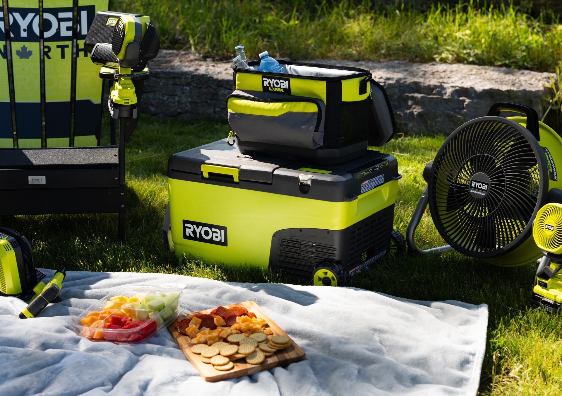 Ryobi cooler, fans, speaker, and picnic food on a blanket in grass.