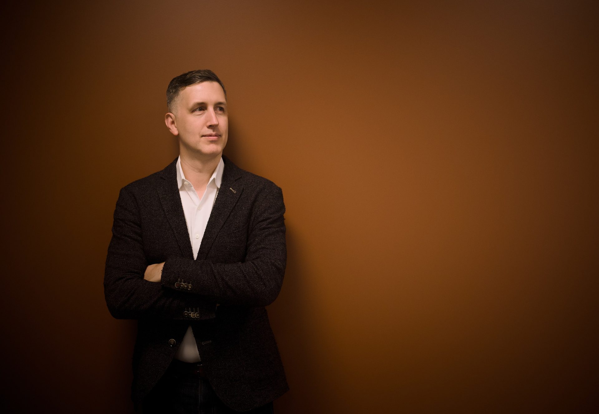 Man in a blazer, arms crossed, looking right, against a brown background.