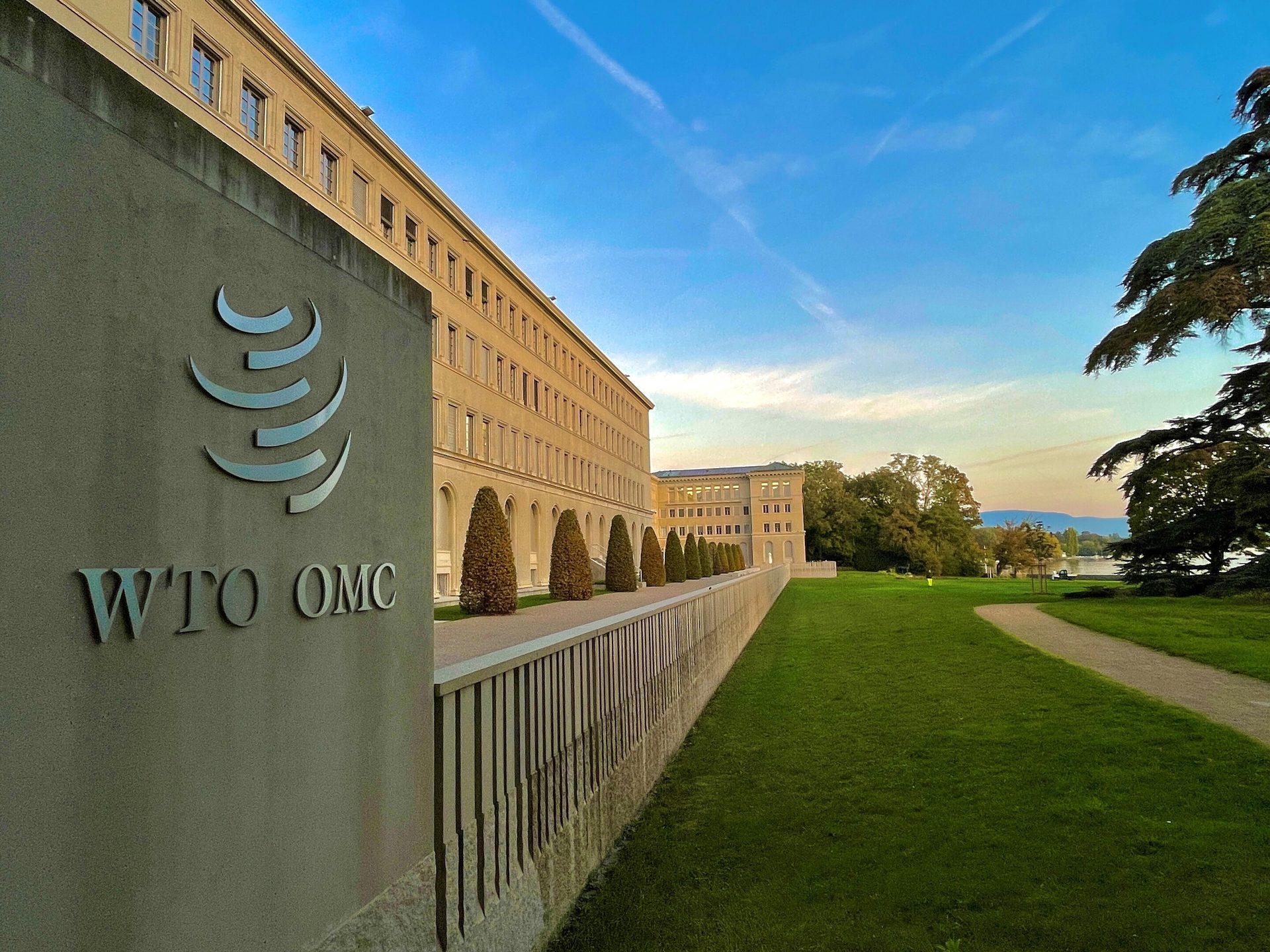 The WTO building in Geneva, Switzerland, featuring its logo on a stone wall, a green lawn, and distant lake and mountains under a blue sky.