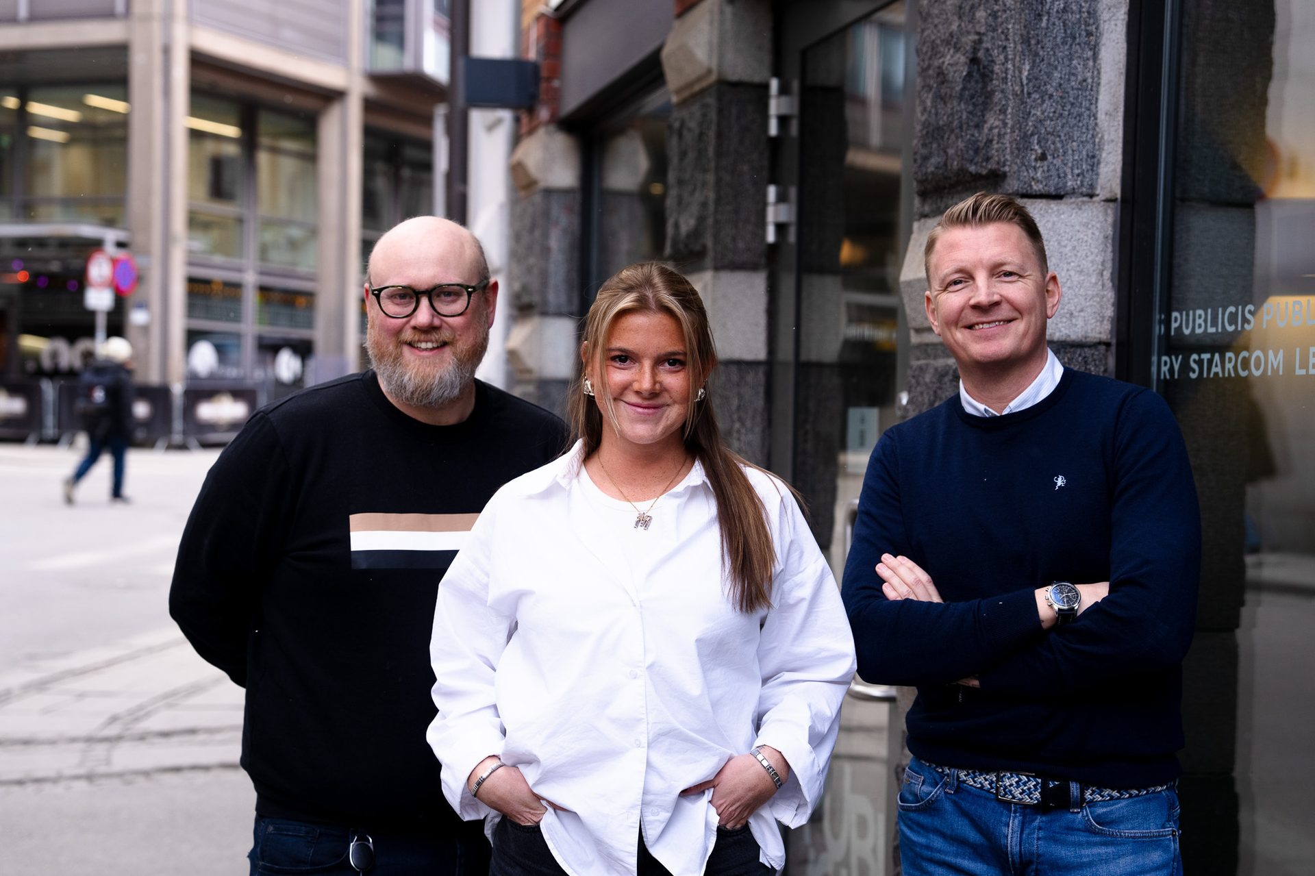 Three smiling colleagues, two men and one woman, stand outdoors in a city setting.