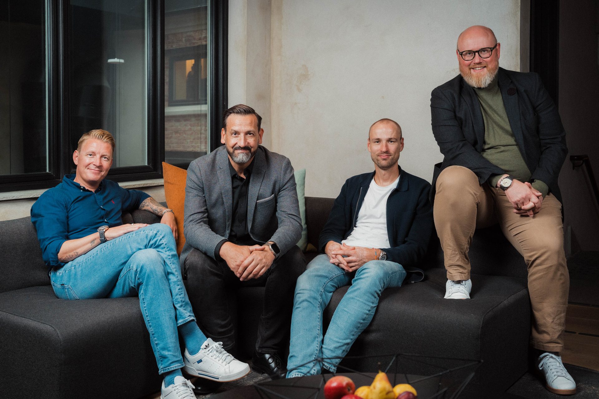 Four smiling men sit on a couch, looking at the camera.