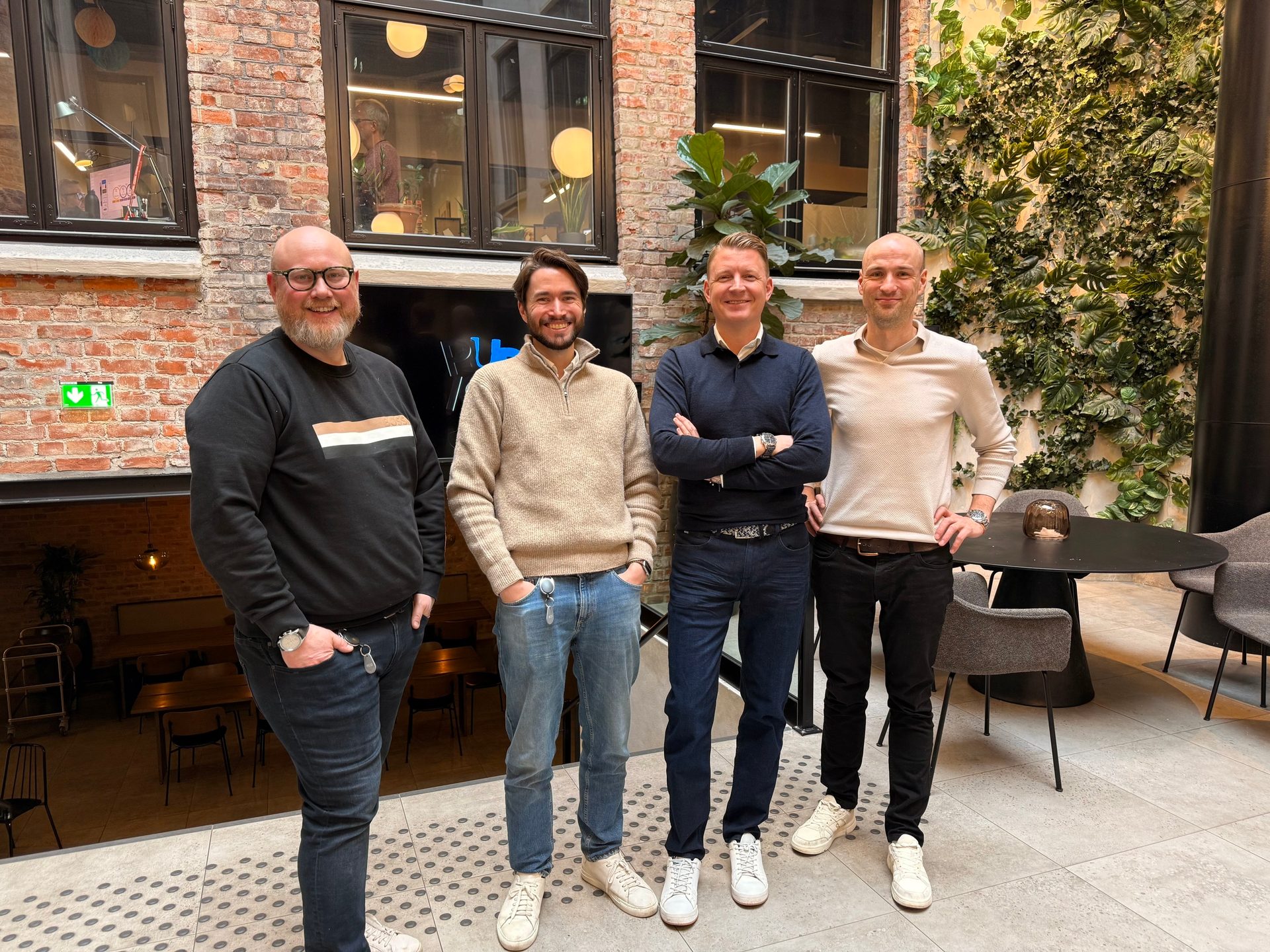 Four smiling men in sweaters and jeans stand in an indoor brick and plant-decorated setting.