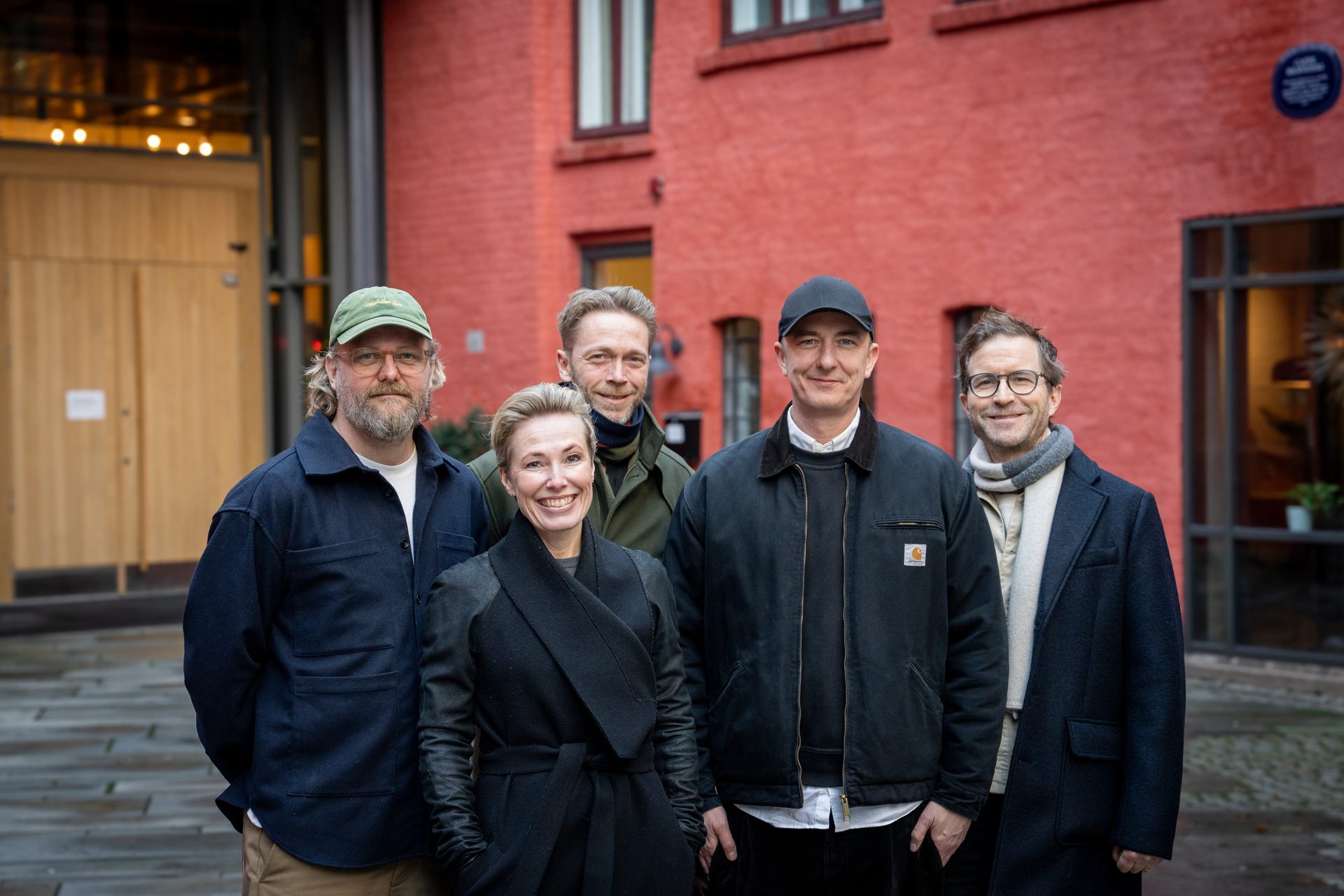 Five people (four men, one woman) smiling in front of a red building.