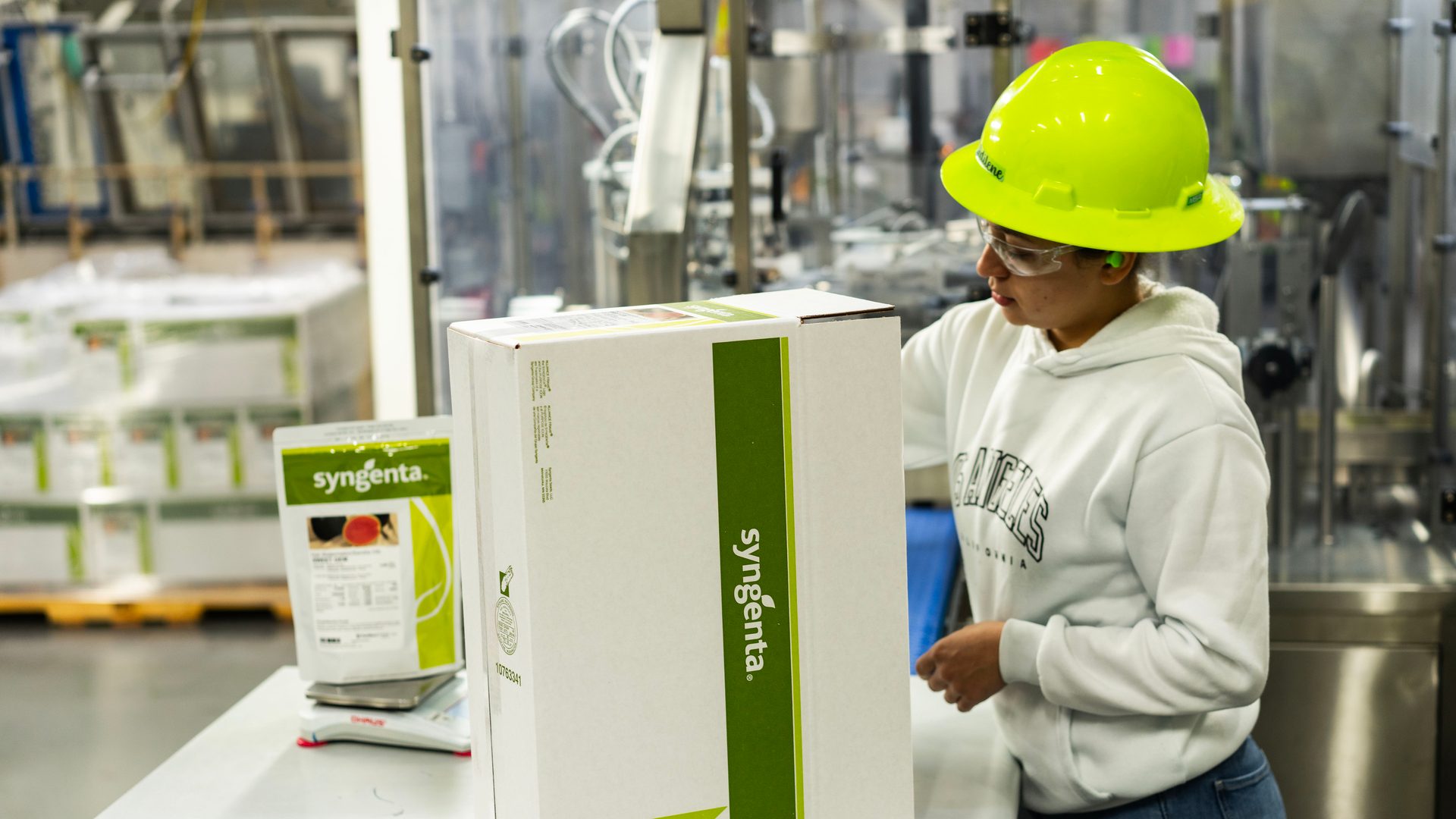 Worker in a hard hat handles a Syngenta product box in a factory, with another package on a scale.