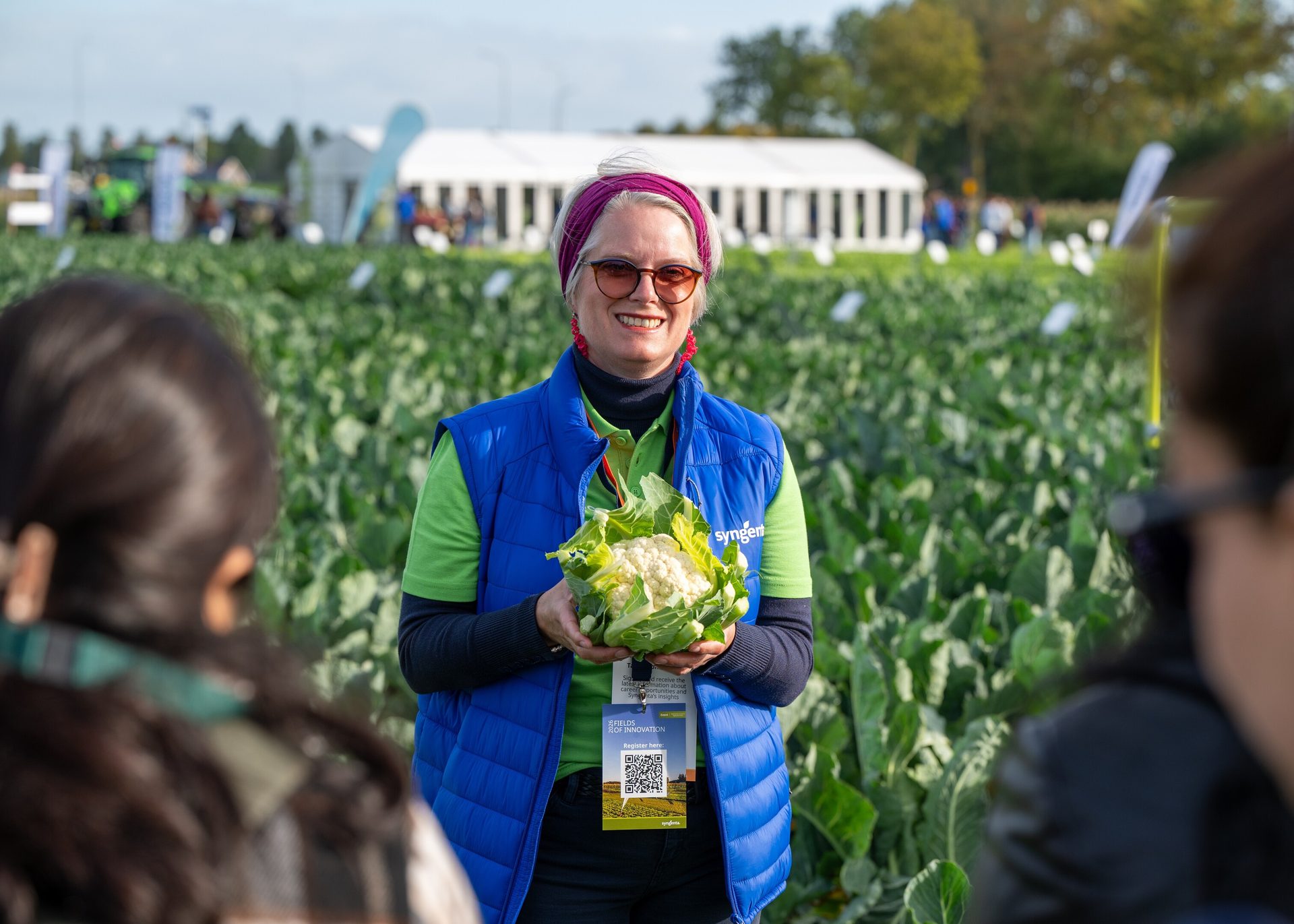 A smiling woman in a blue vest holding a cauliflower in a field, with tents in the background.