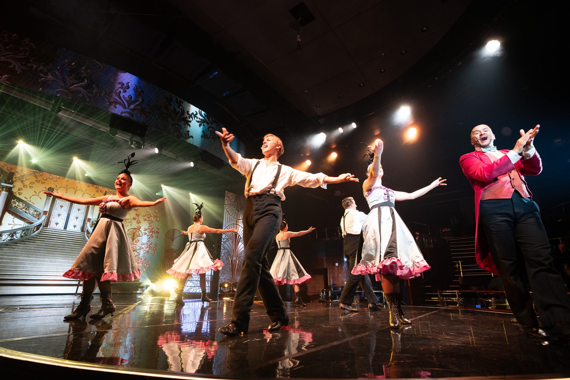 Performers in period costumes dance on a reflective stage under bright spotlights.