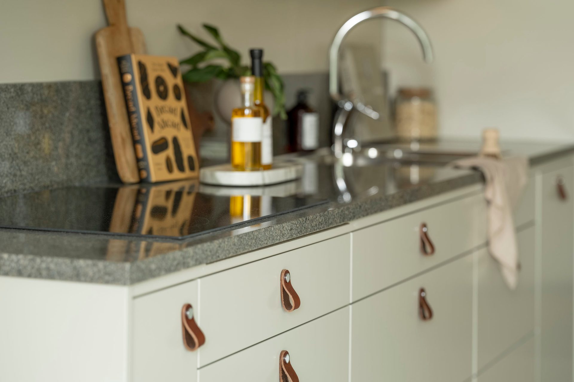 Kitchen counter with cooktop, sink, cookbook, oil bottles, and cabinets with leather handles.