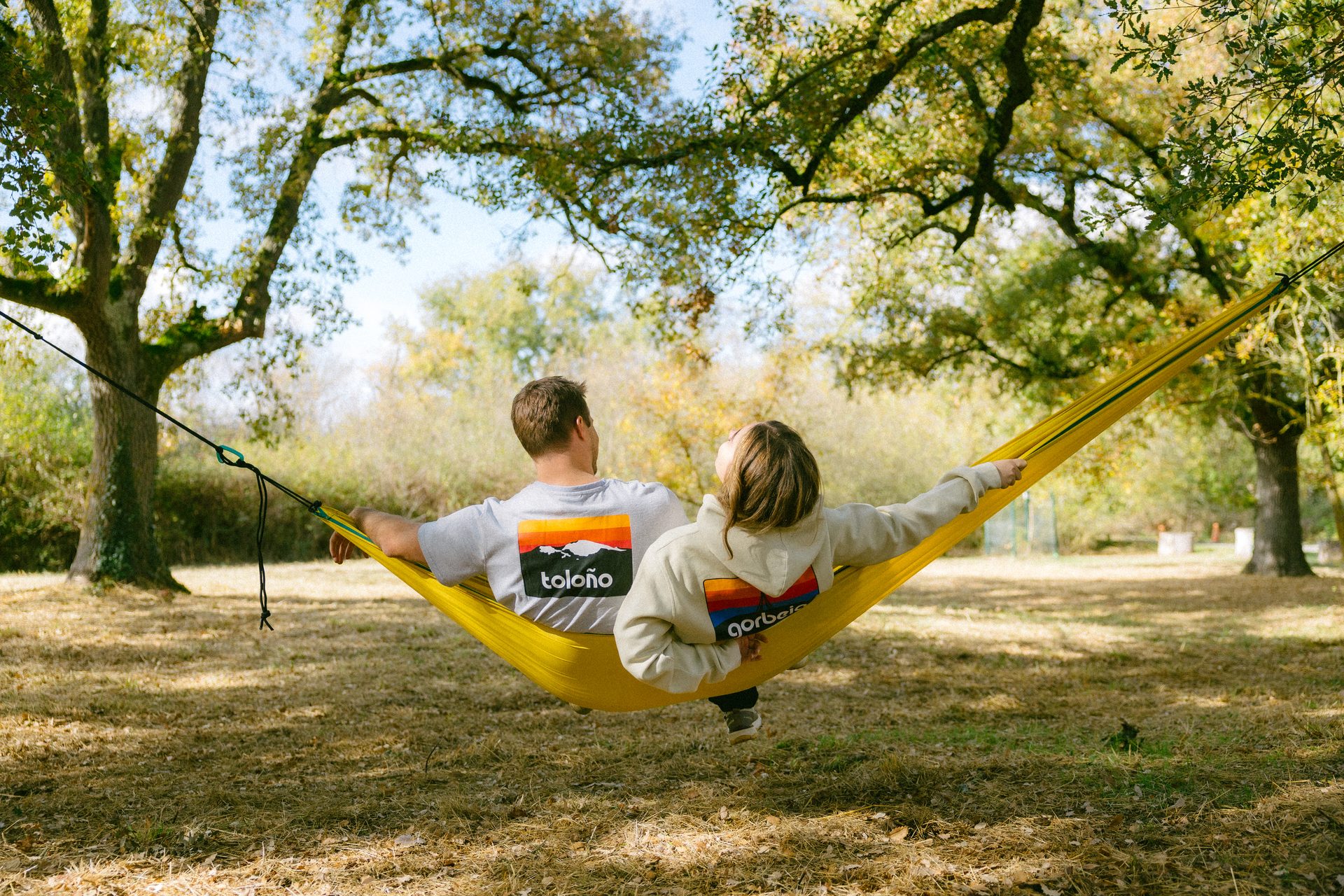 Two people relax in a yellow hammock outdoors, backs to camera, looking up at trees.