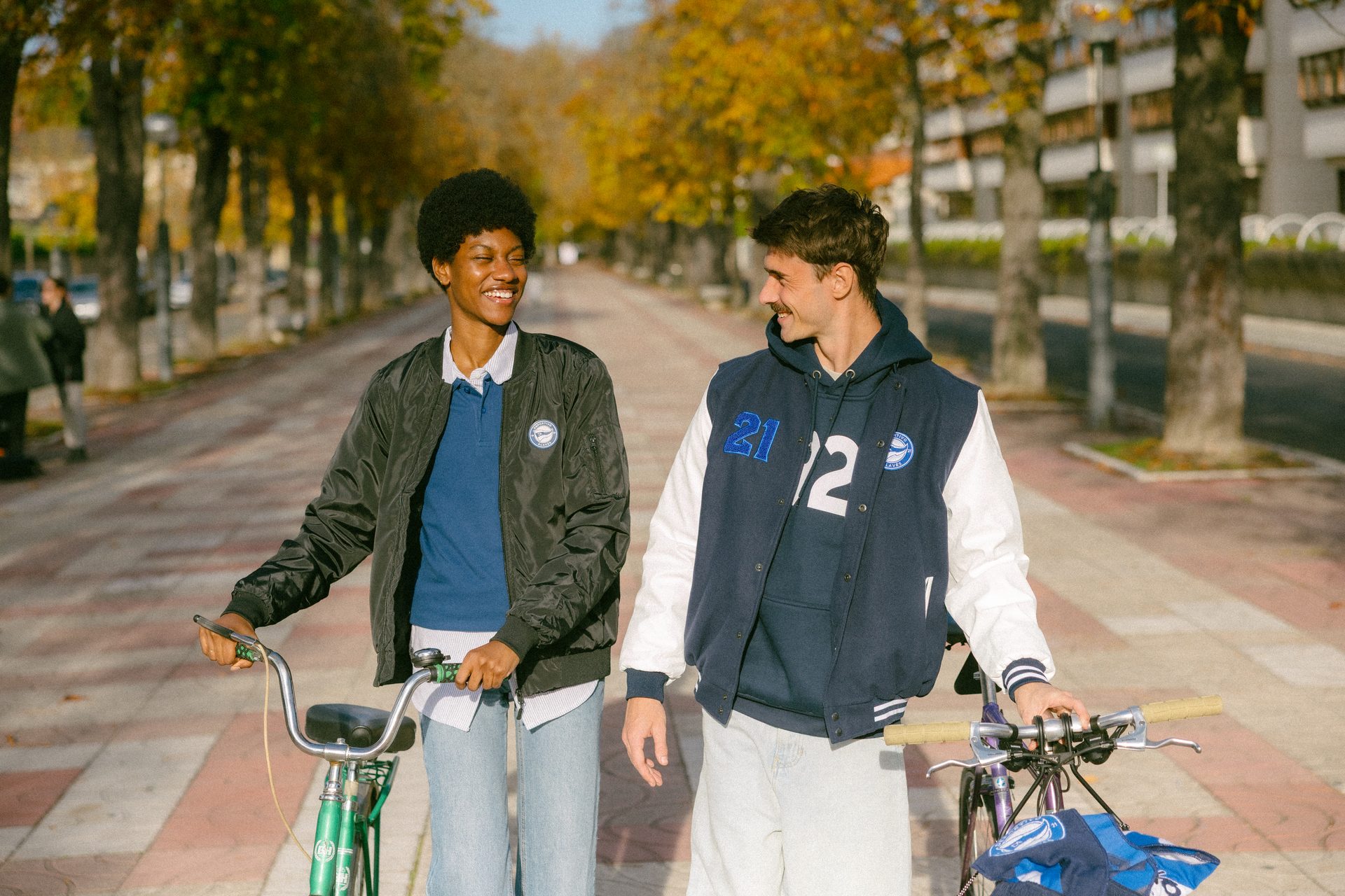Two smiling friends walk with their bicycles on a sunny, autumn street.