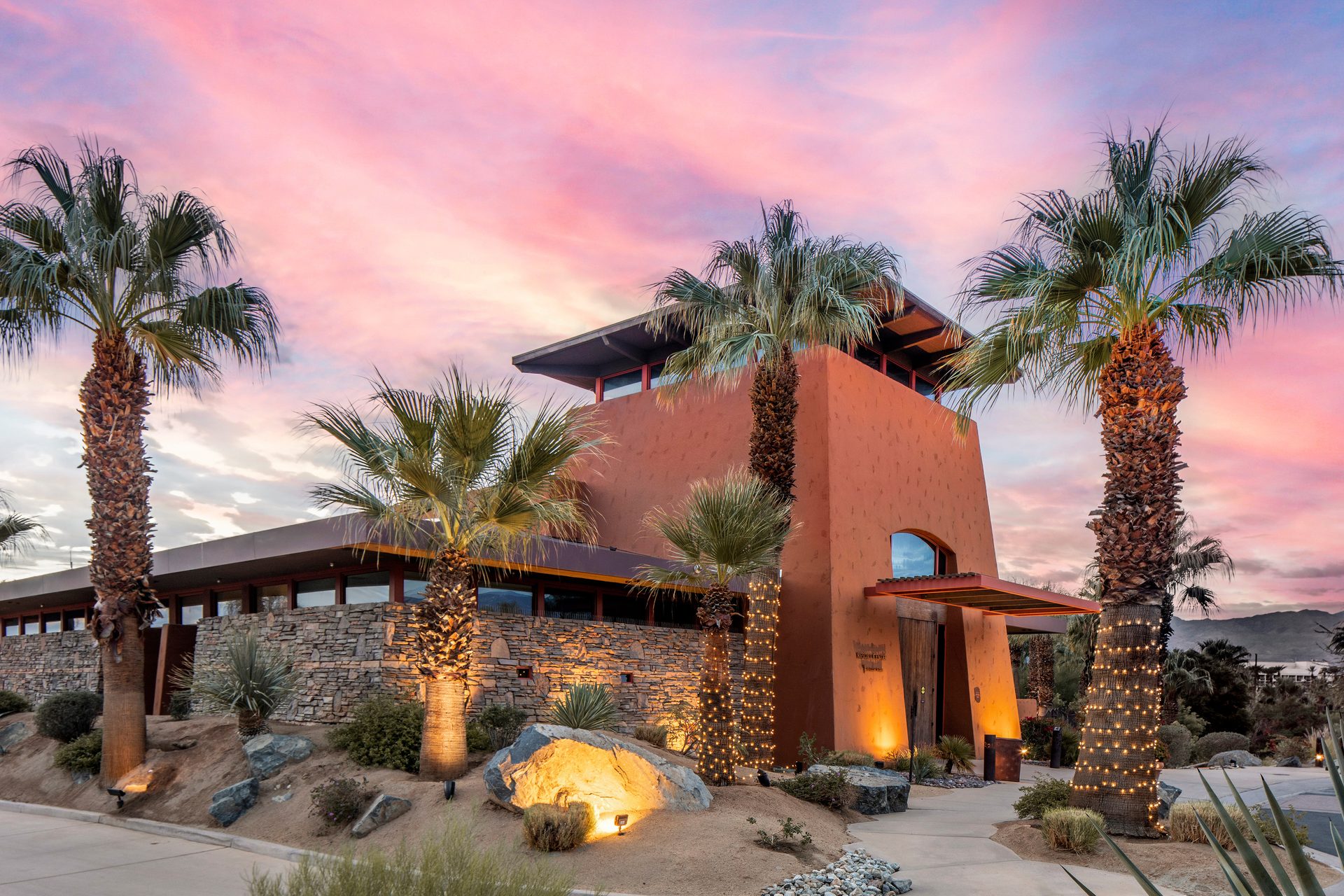 Terracotta building with stone, lit palm trees, and a vibrant pink-purple sunset.