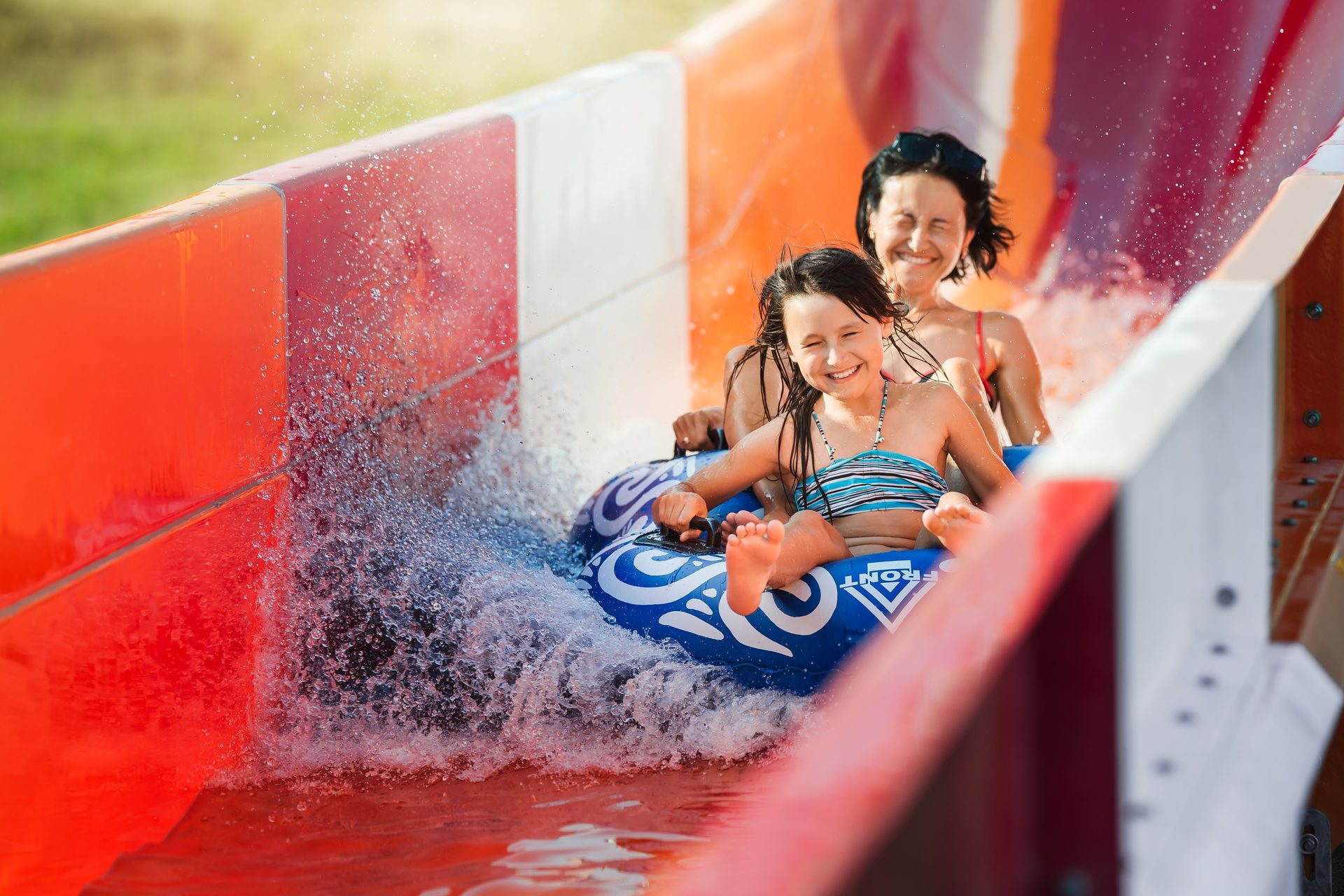 A mother and daughter joyfully ride a water slide on a blue tube, splashing water.